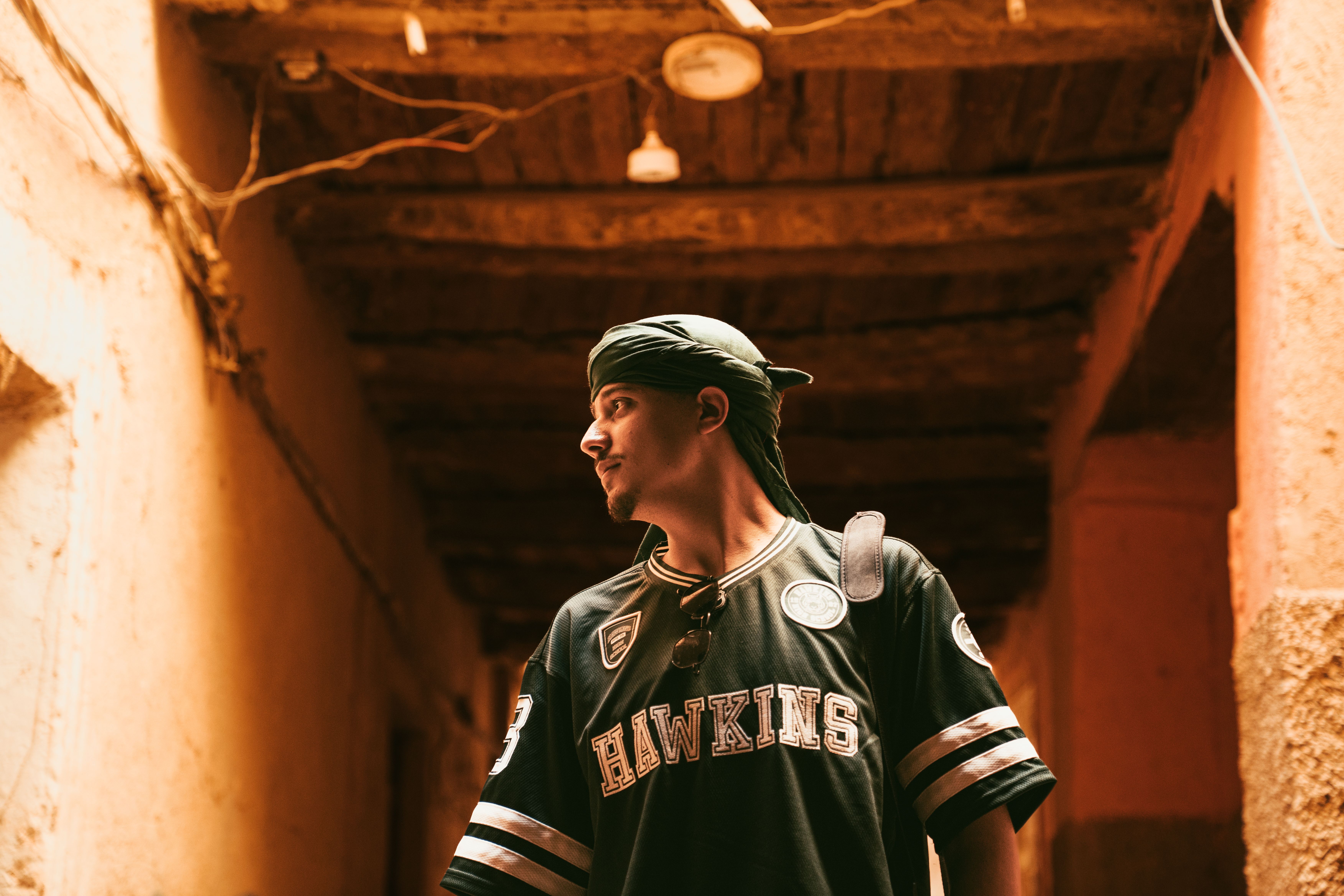 A young man wearing a bandana explores a rustic, dimly lit alleyway.