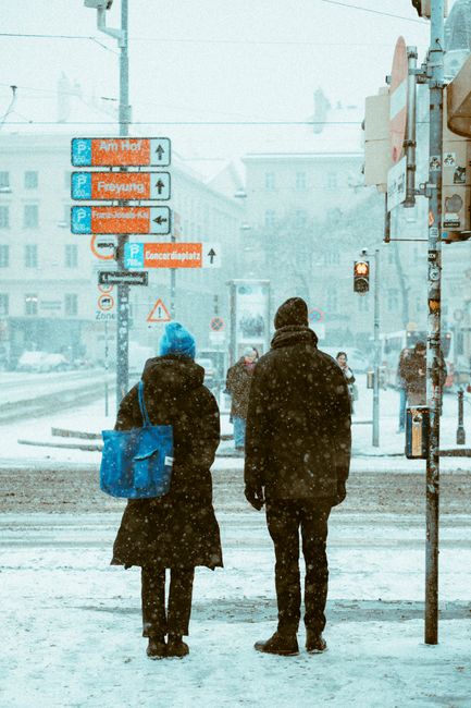 Two people stand on a snowy street with directional signs in a city setting.