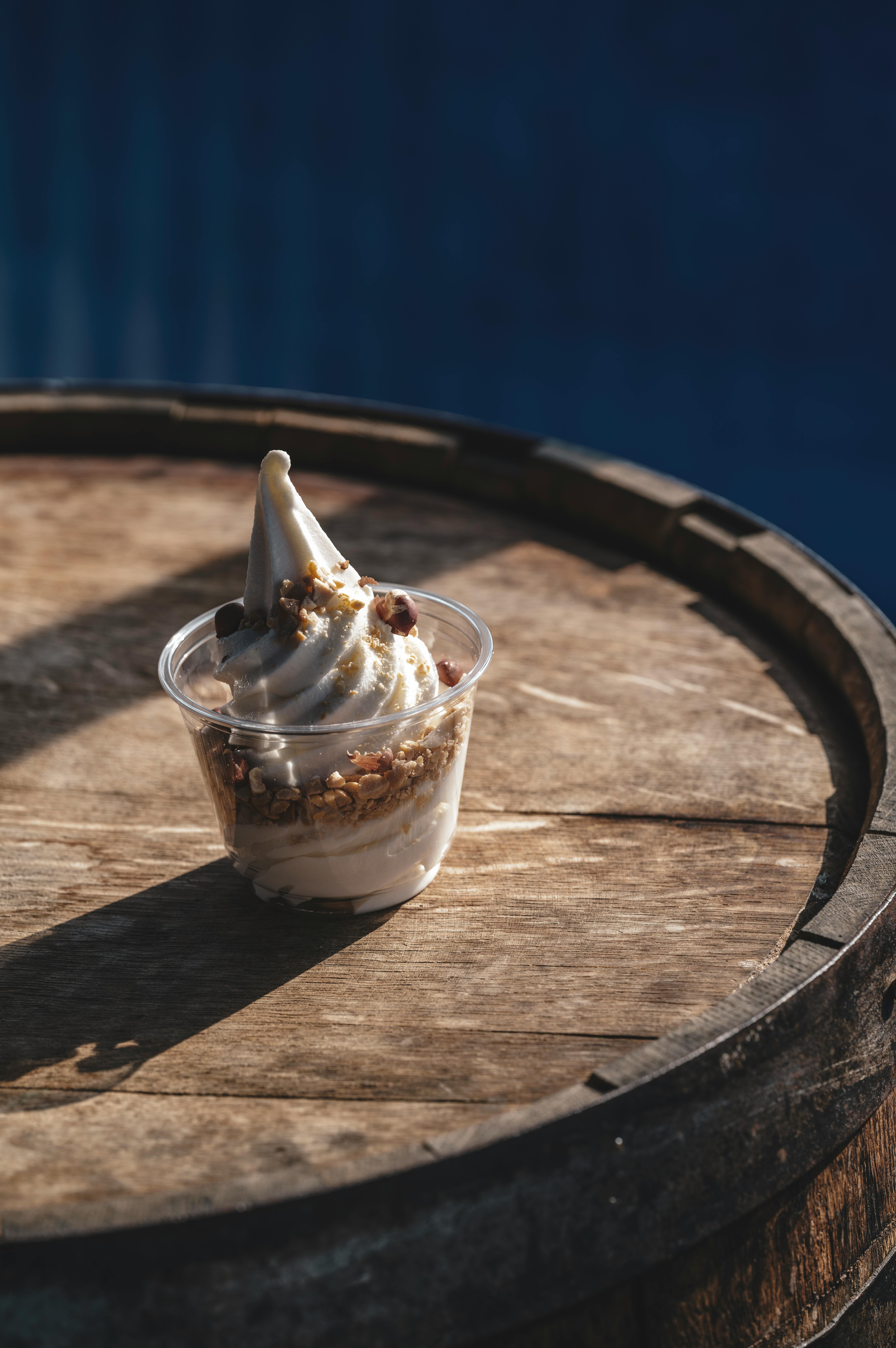 A cup of soft serve ice cream with toppings on a wooden barrel in sunlight.