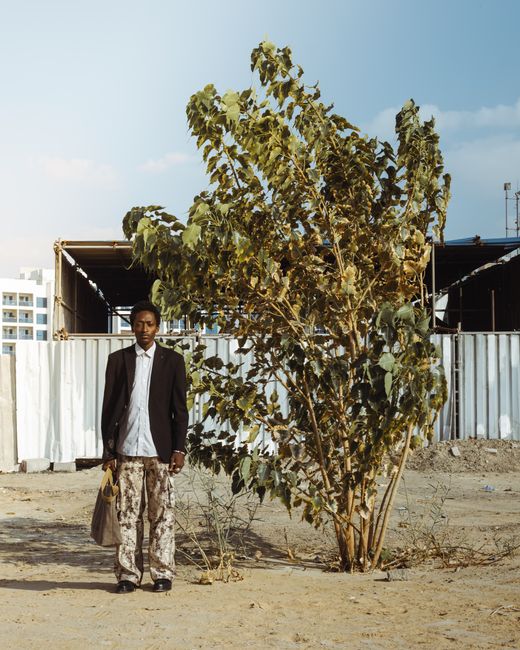 Man stands by tree in an urban area, carrying a bag, against construction backdrop.