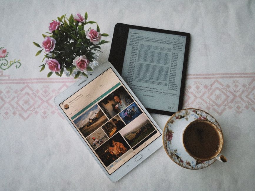 A cozy flat lay featuring a tablet, e-reader, decorative flowers, and a cup of coffee on a patterned cloth.