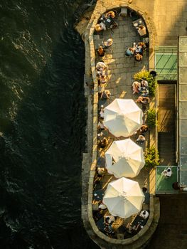 Aerial shot of an outdoor cafe by the river at sunset with patrons enjoying their time under umbrellas.