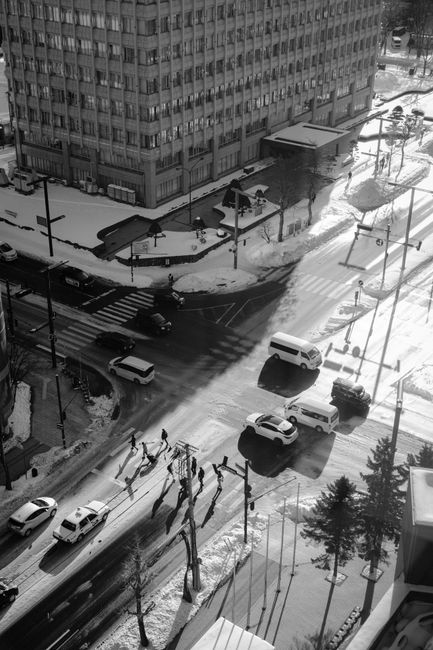 Aerial black and white image of a snowy urban intersection with pedestrians and vehicles in city winter setting.