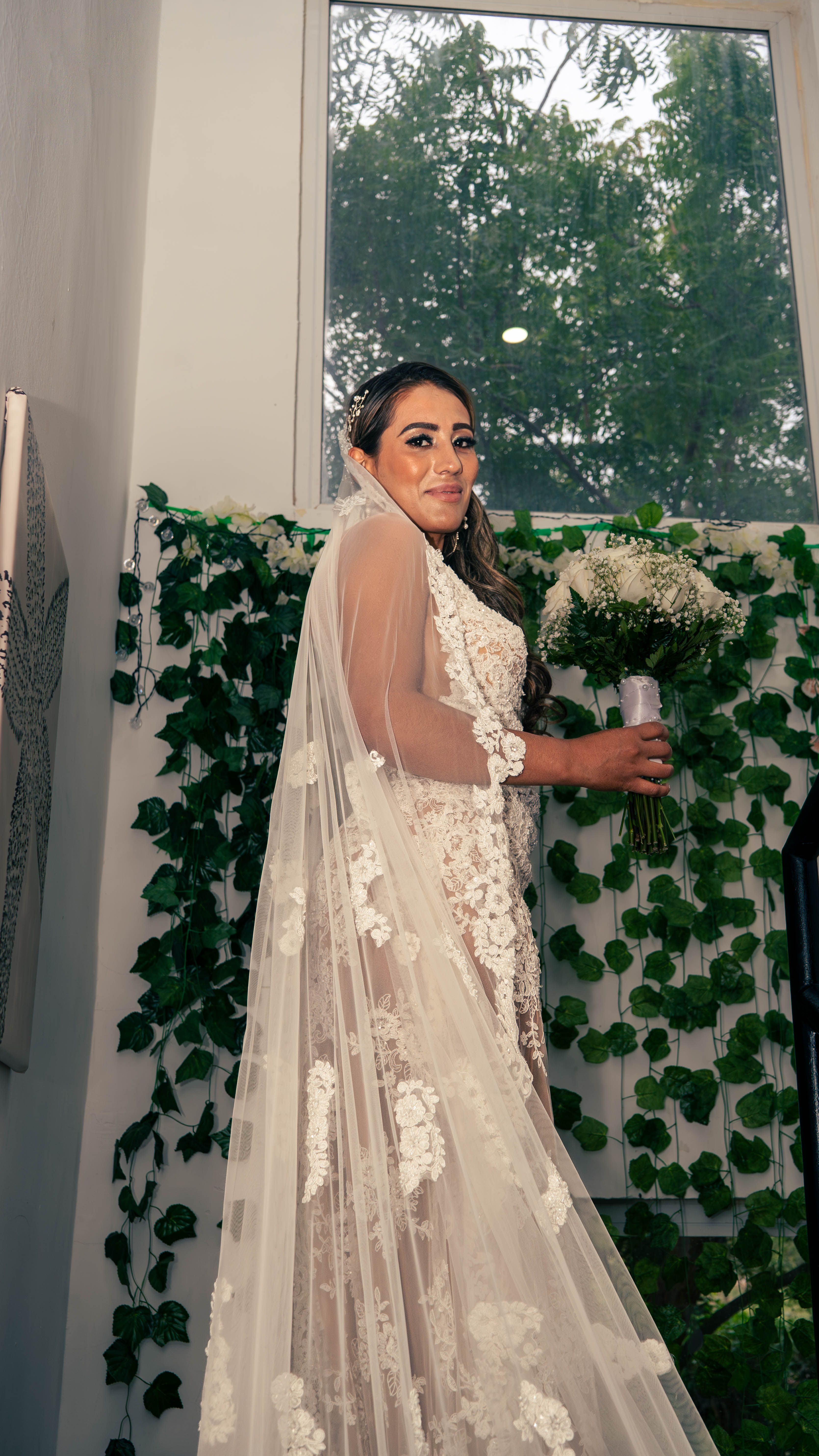 Bride in a lace dress holding a bouquet, captured indoors with greenery backdrop.