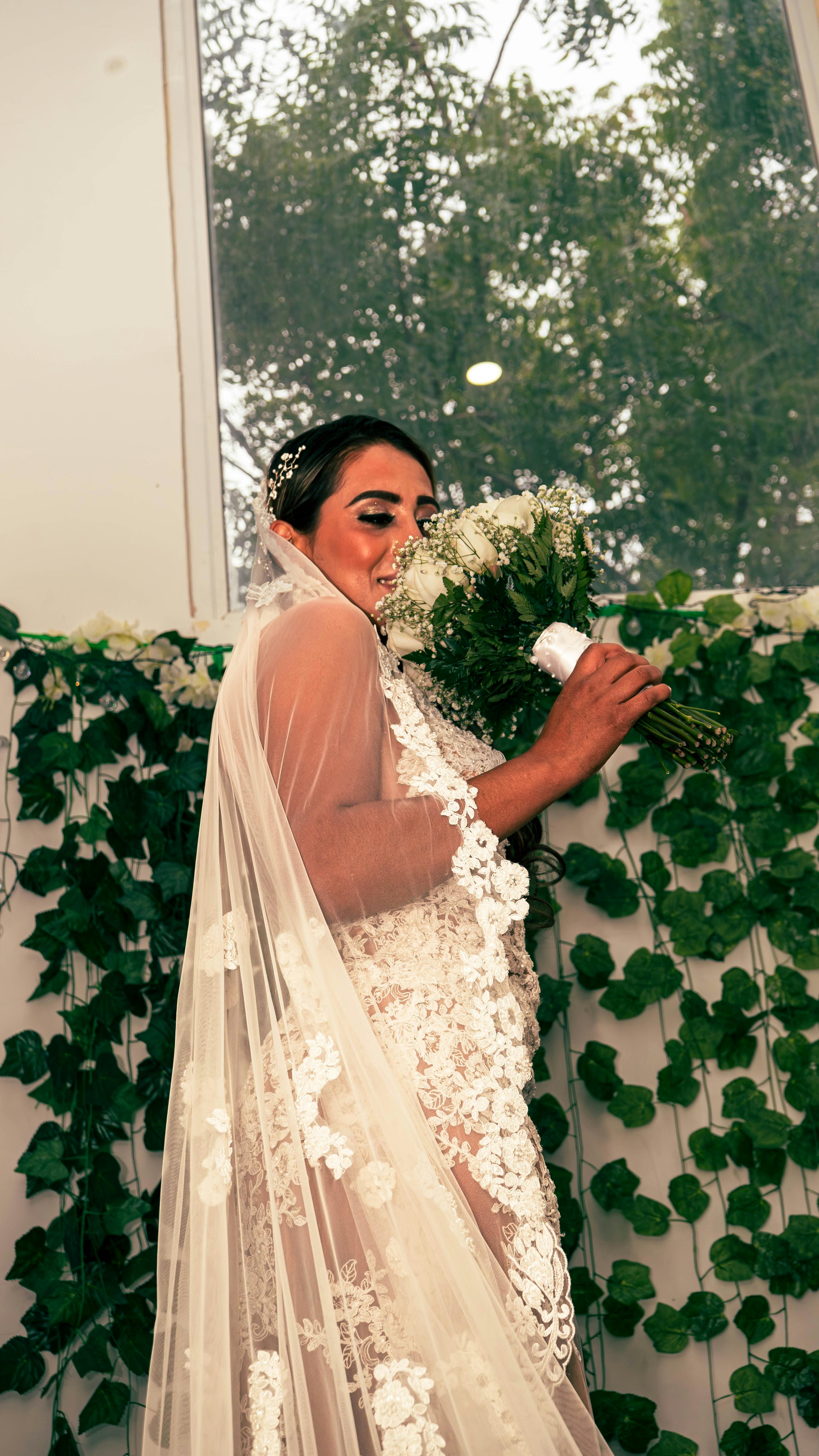 An elegant bride in a lace gown holding a bouquet amidst floral decorations indoors.