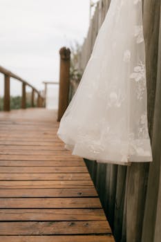 Delicate white wedding dress with floral patterns hanging by a rustic wooden pathway, capturing a romantic outdoor setting.