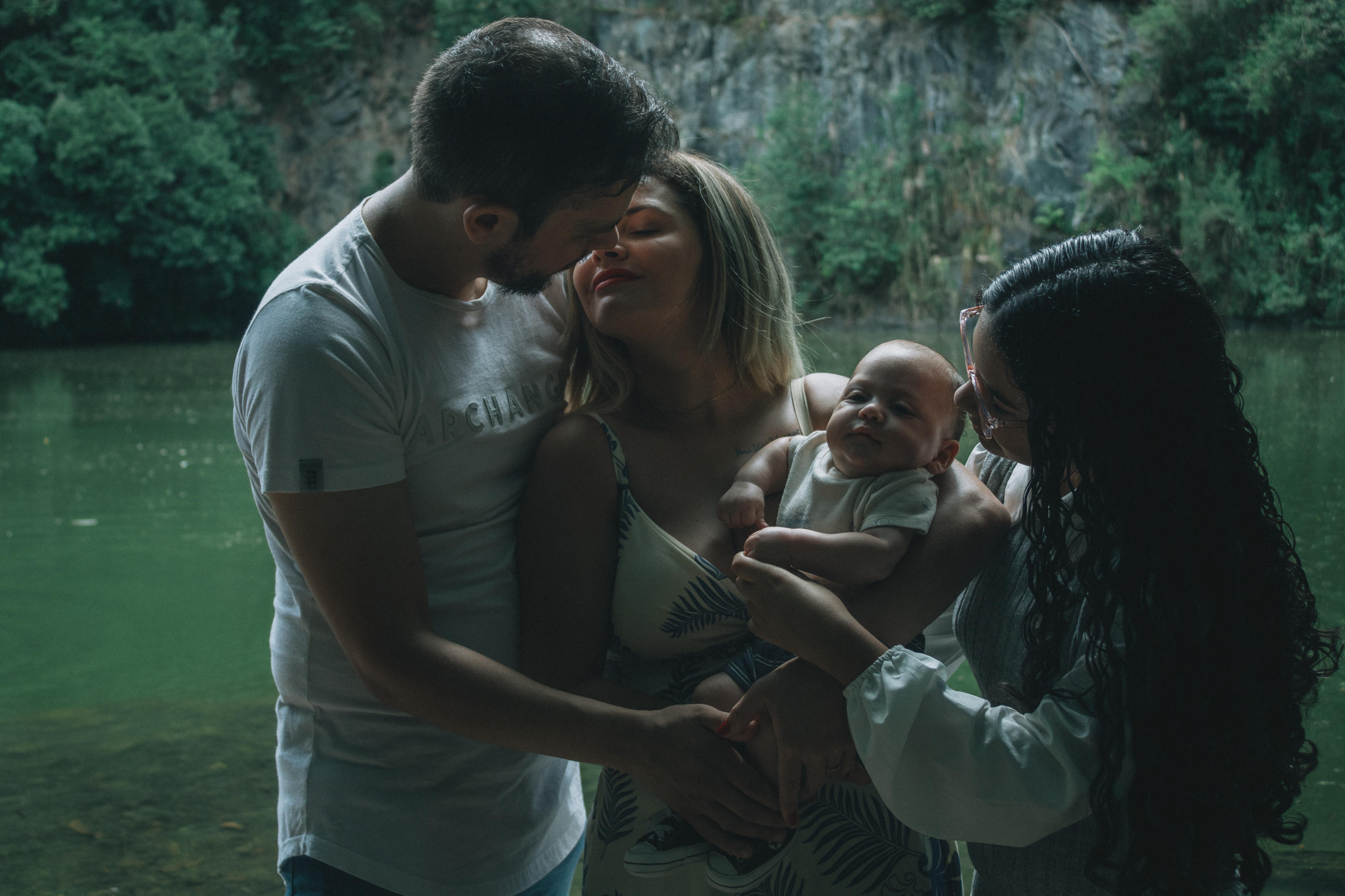 ฟรี ครอบครัวหนึ่งกำลังเพลิดเพลินกับช่วงเวลาอันแสนอบอุ่นร่วมกันริมทะเลสาบอันเงียบสงบที่รายล้อมไปด้วยพืชพรรณเขียวชอุ่ม คลังภาพถ่าย