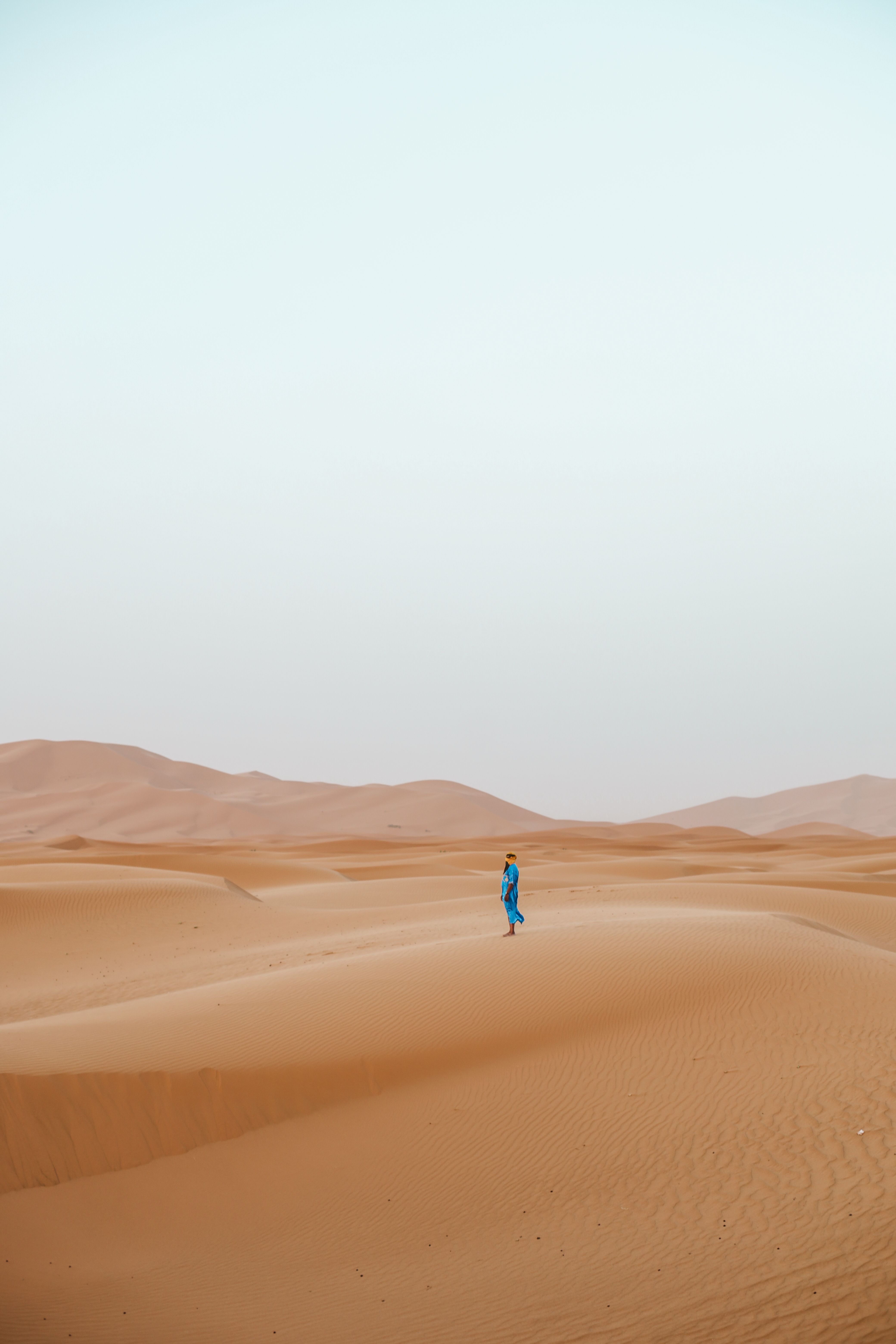 A lone person in blue clothing walks across the vast dunes of the Sahara Desert in Morocco.