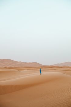 A lone person in blue clothing walks across the vast dunes of the Sahara Desert in Morocco.