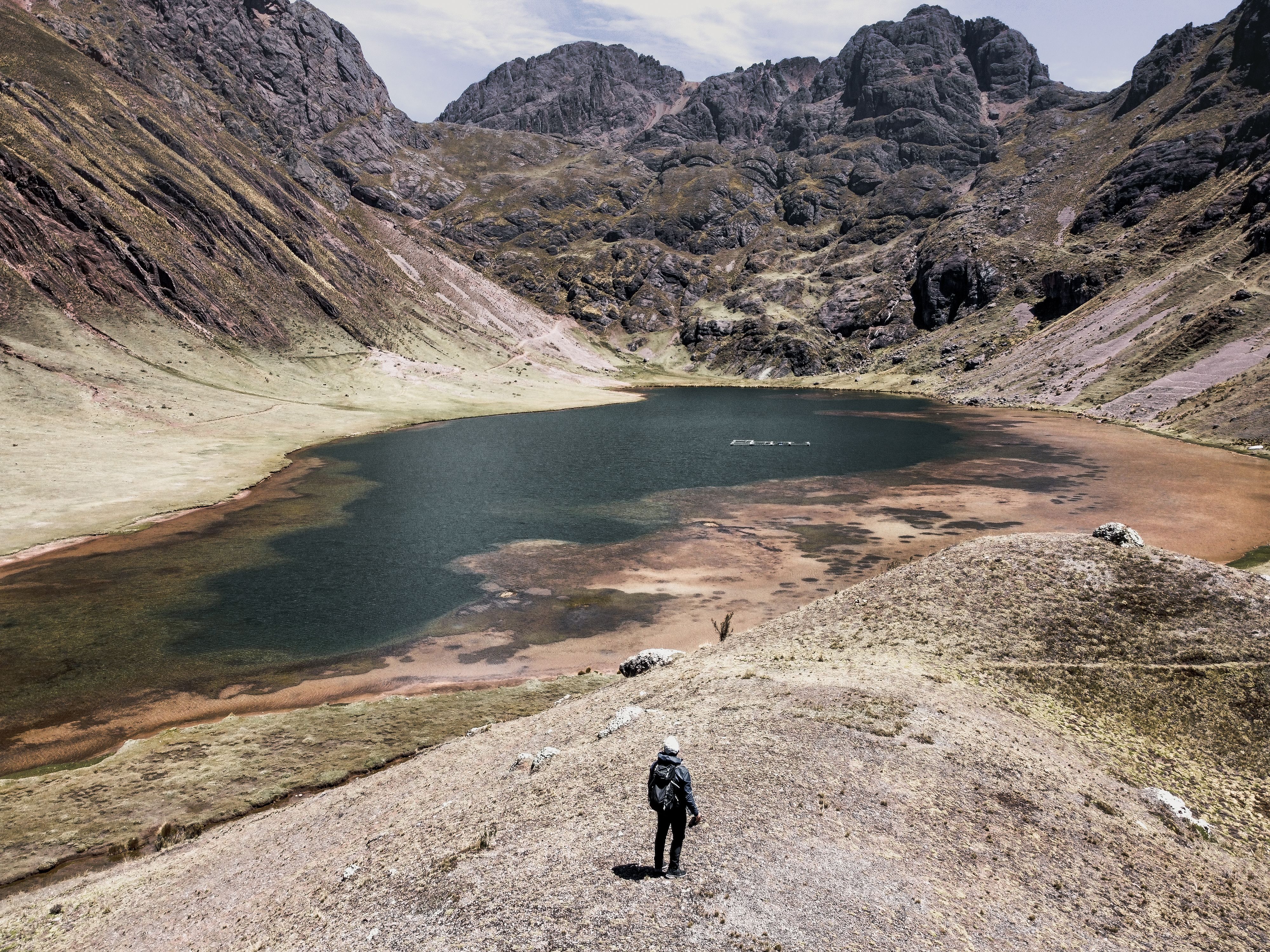 A lone hiker stands on a mountain slope, admiring a remote lake nestled between rugged peaks.