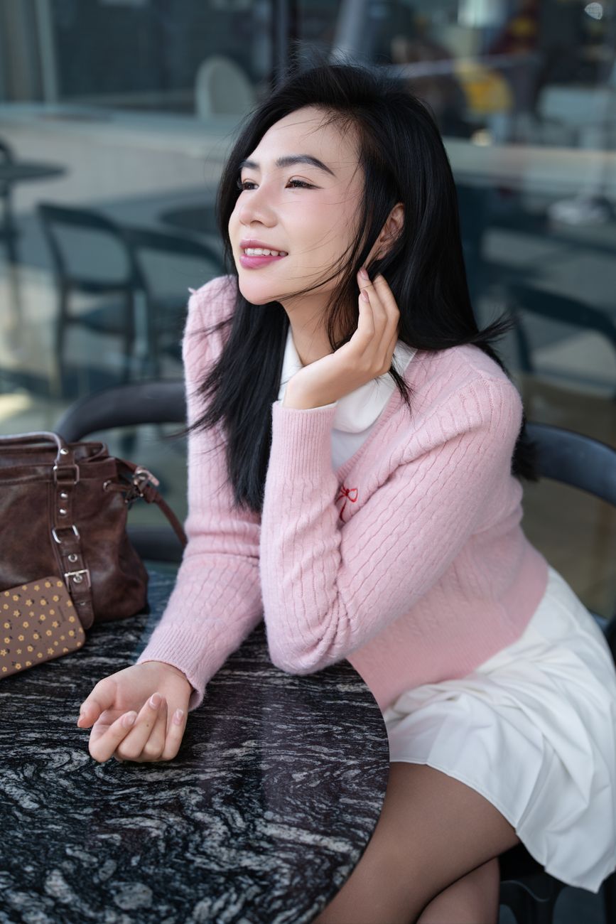 Young woman in a pink sweater sitting at an outdoor cafe, smiling and looking relaxed.