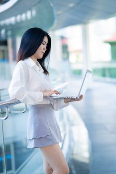 Confident young Asian woman working remotely with a laptop in a modern setting, reflecting digital nomad lifestyle.