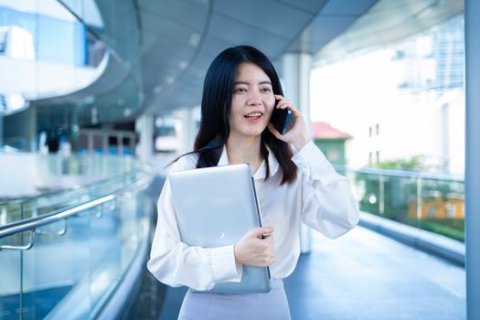 Asian woman in smart casual attire using phone and holding laptop outdoors in Bangkok.