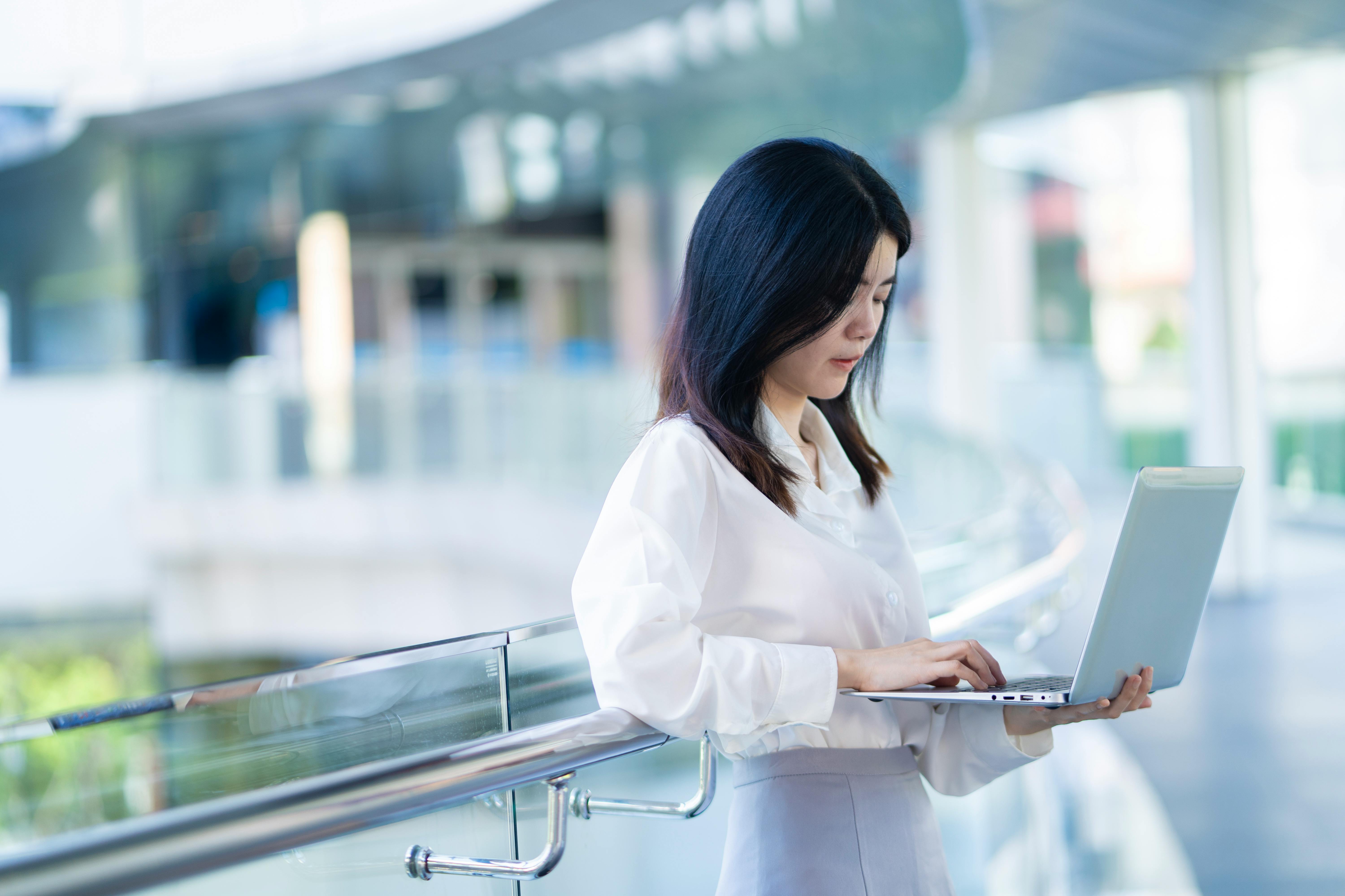 Asian businesswoman multitasks on a laptop in a modern office setting.