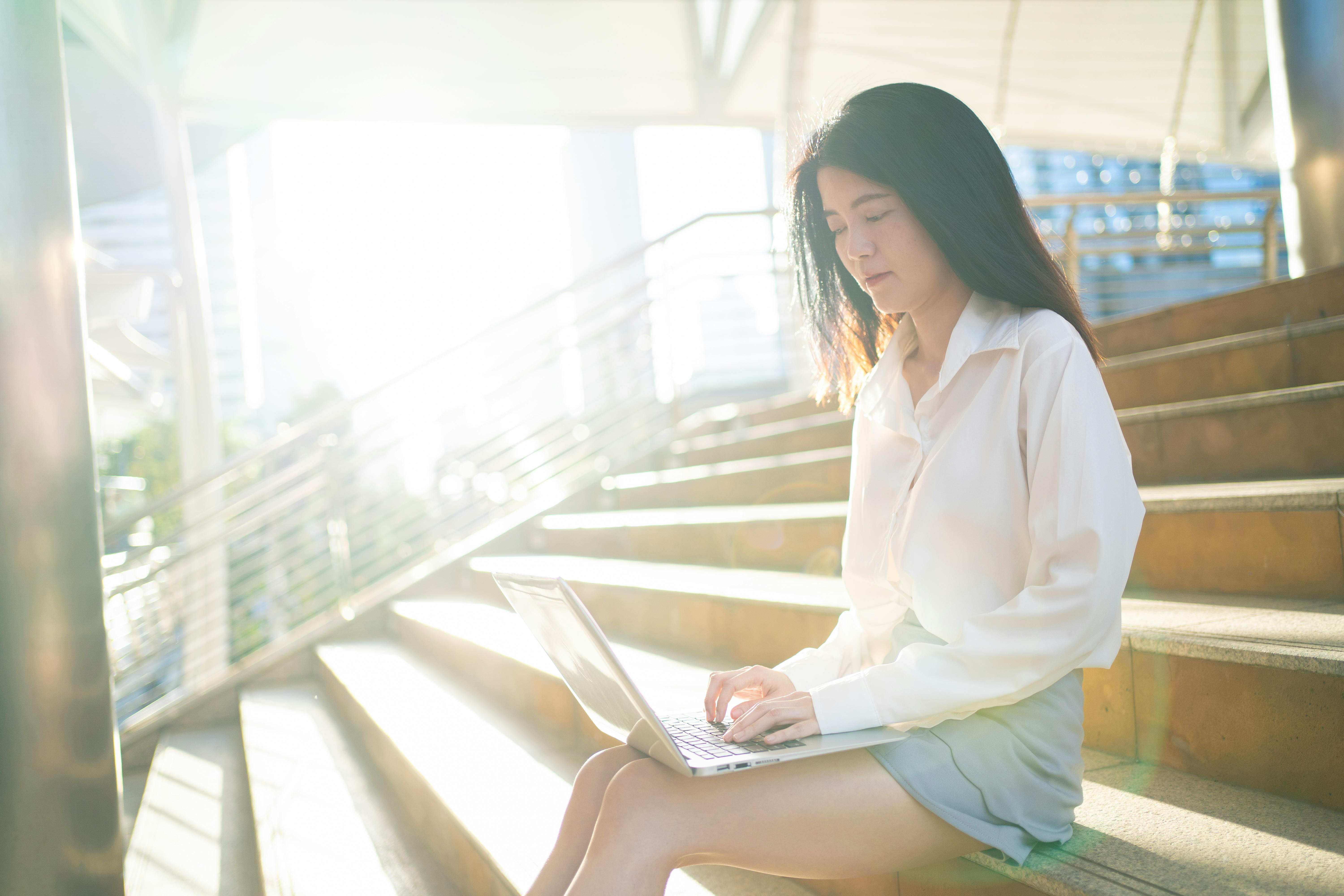 Young Asian woman working remotely on stairs with laptop, sunny urban setting in Bangkok.