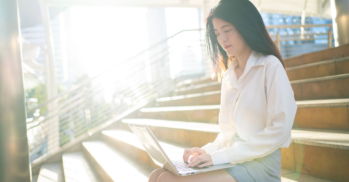 Young Asian woman working remotely on stairs with laptop, sunny urban setting in Bangkok.