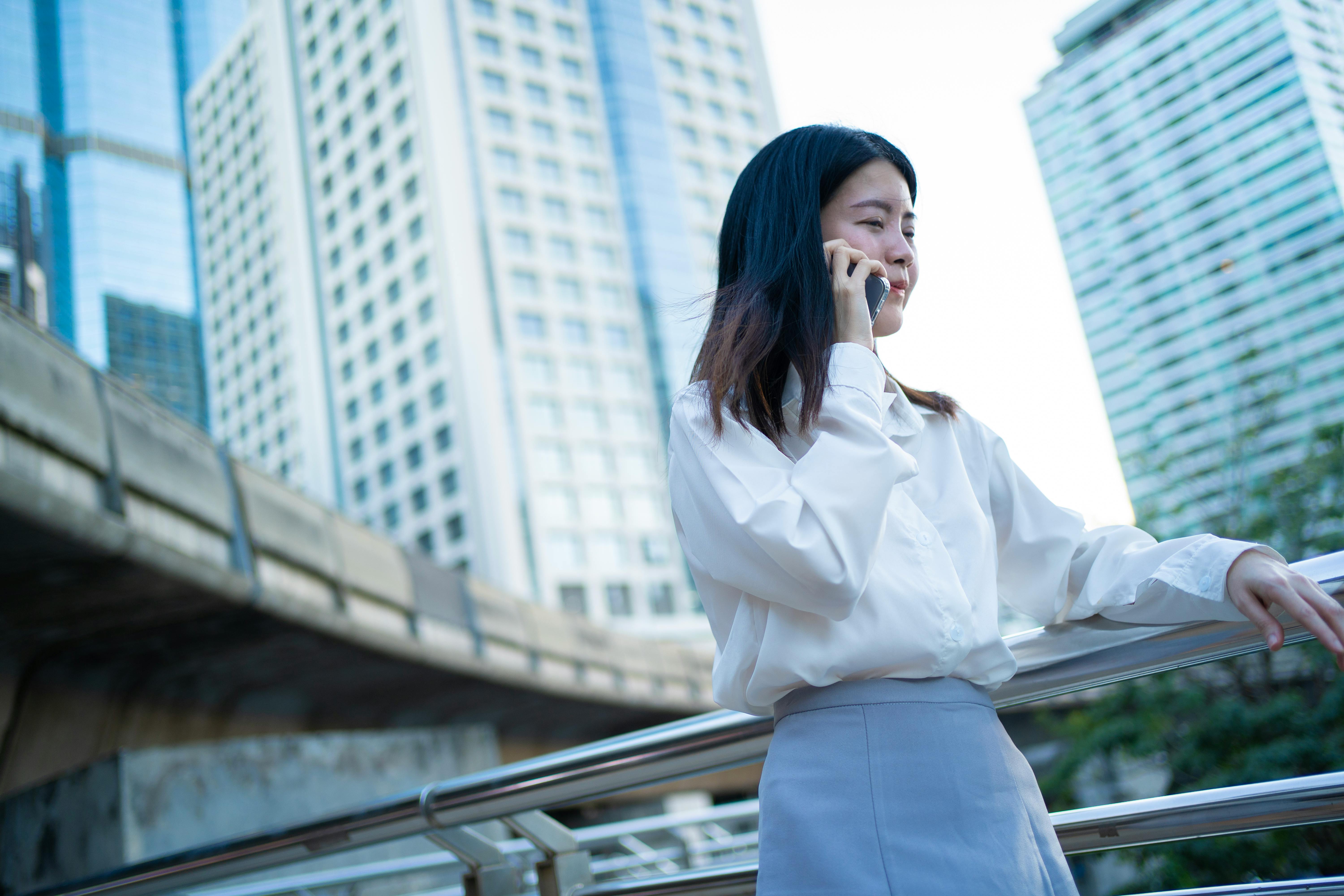 Confident Asian woman in business attire talks on phone in Bangkok's cityscape.