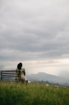 A woman sitting alone on a bench, gazing at distant mountains under a cloudy sky.