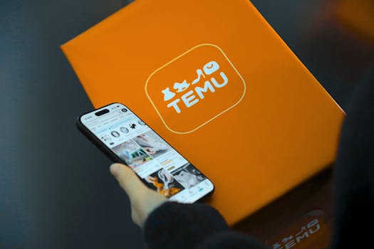 Hand holding smartphone with Temu app beside orange Temu package.