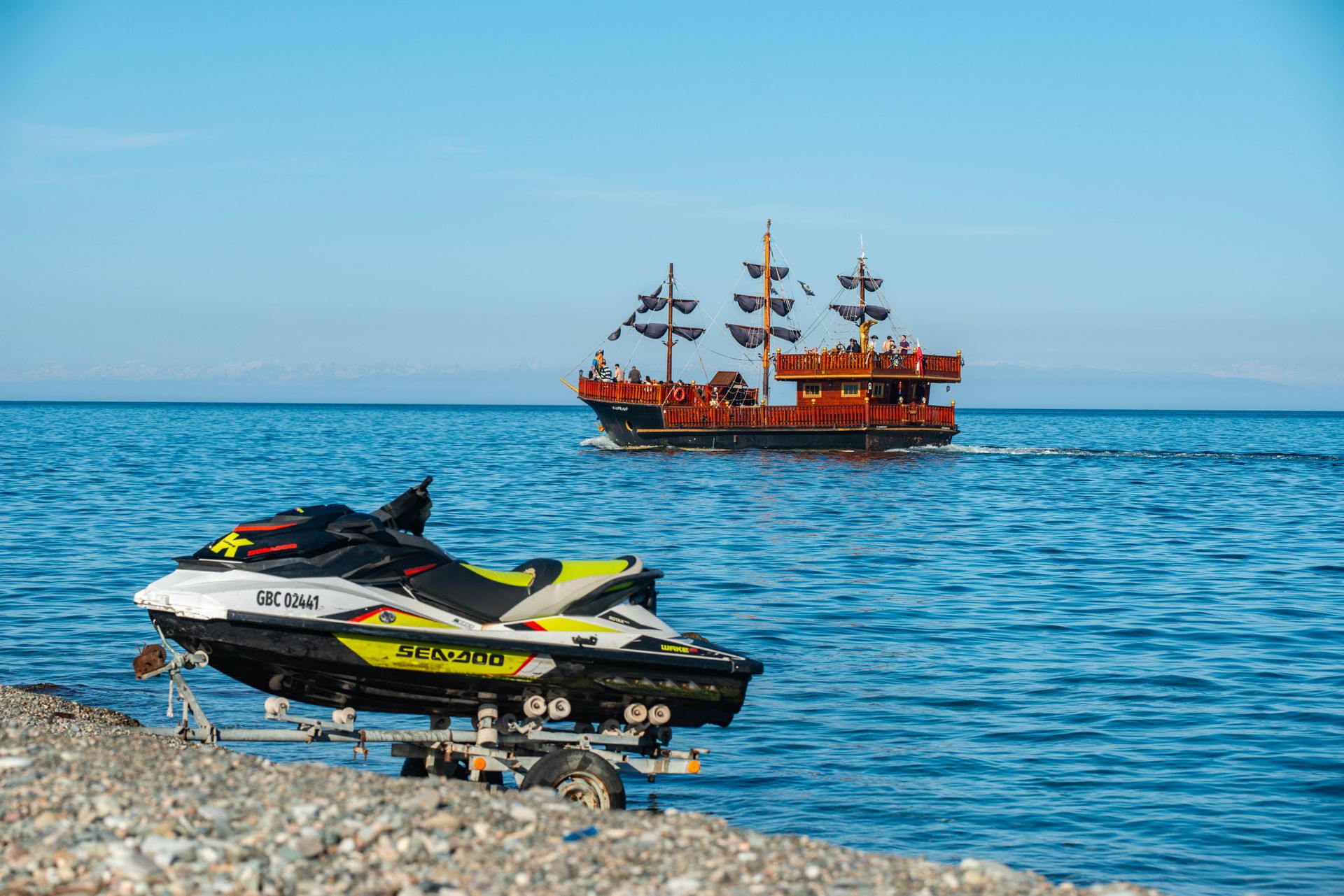 A jet ski on a pebble beach with a pirate ship sailing in the sunny ocean.