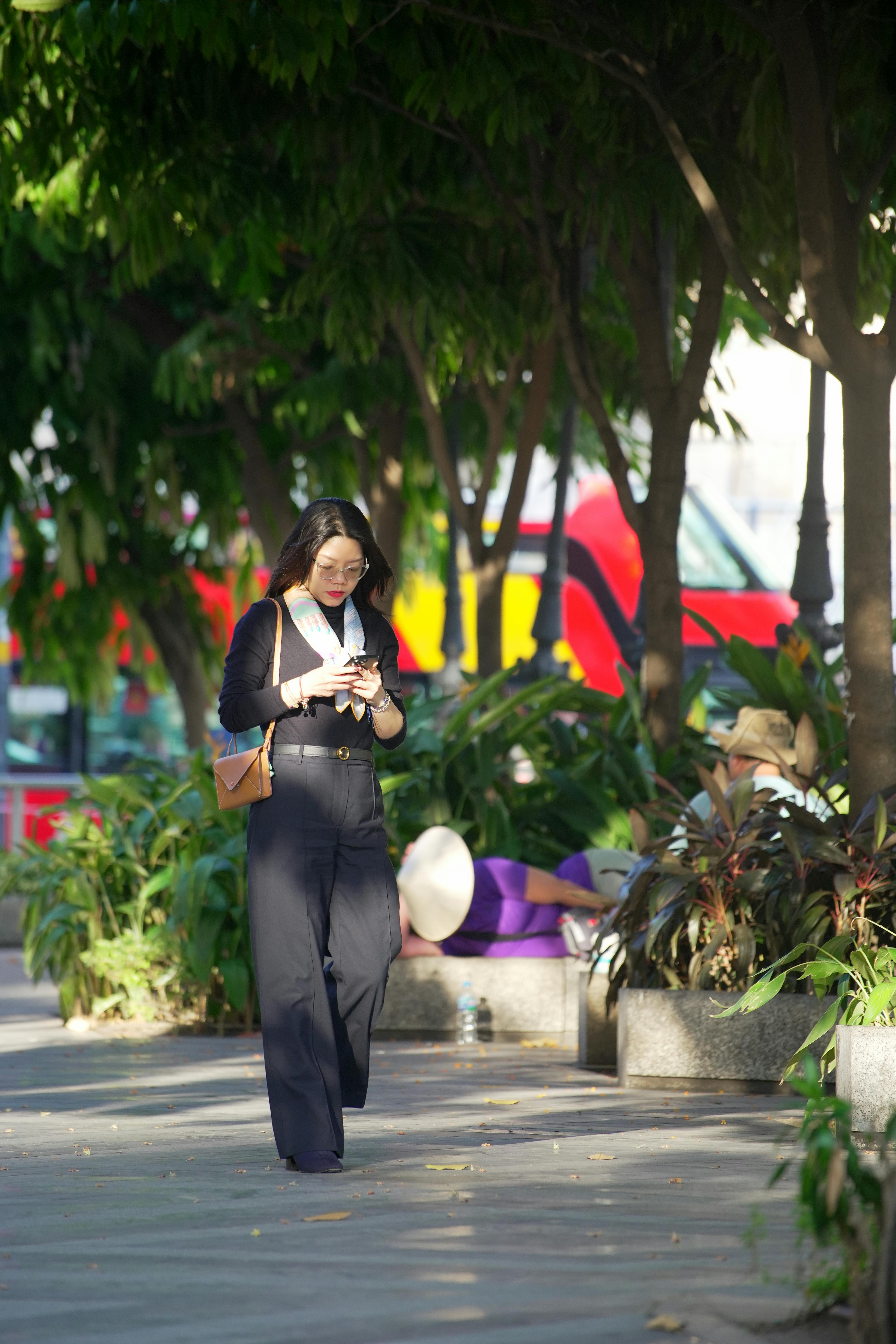 De franc Dona asiàtica passejant per un parc de la ciutat mentre utilitza el seu telèfon intel·ligent. Arbres il·luminats pel sol i autobús urbà al fons. Foto d'estoc
