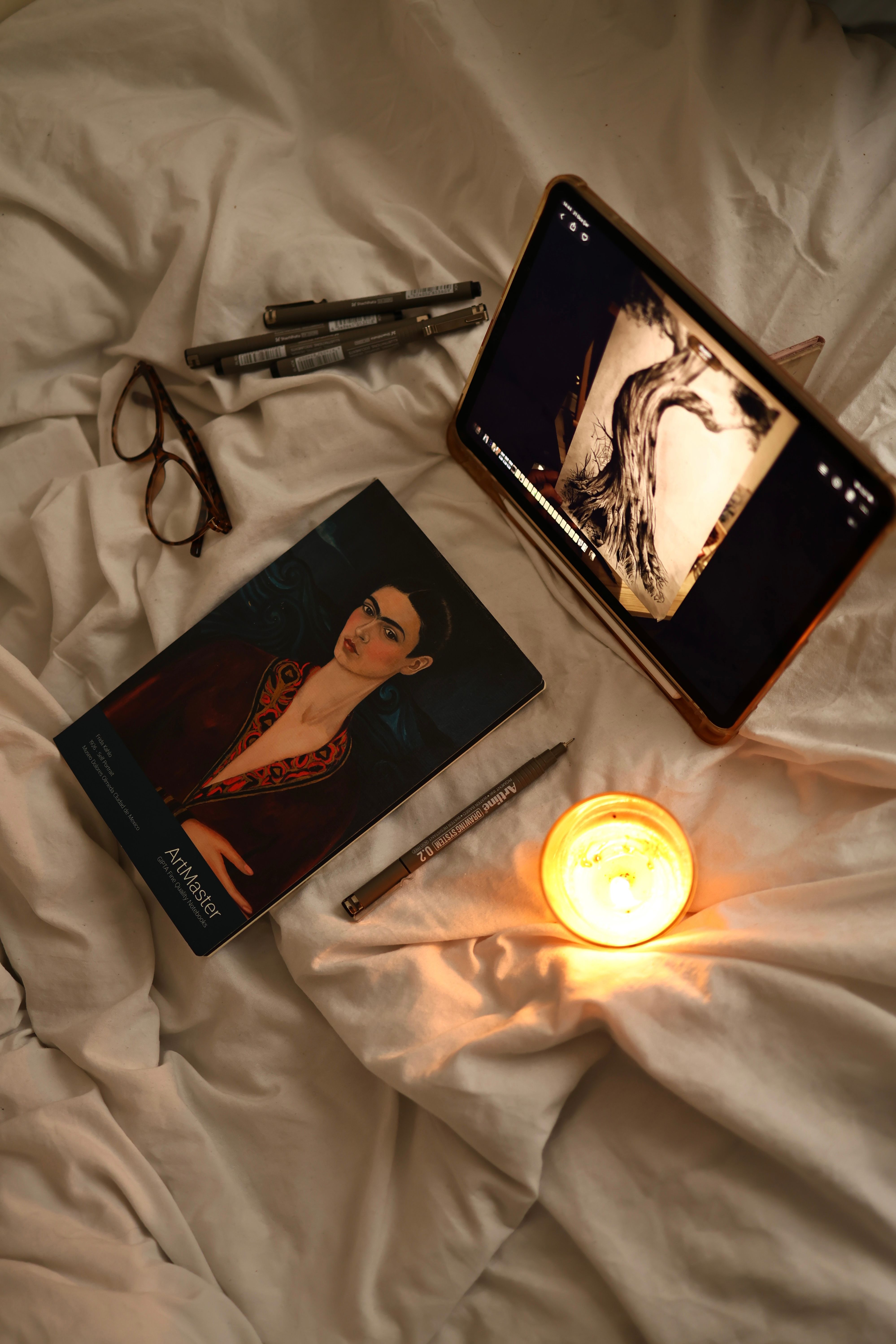 An artistic setup featuring a tablet displaying art, book, and candle on a cozy bed setting.