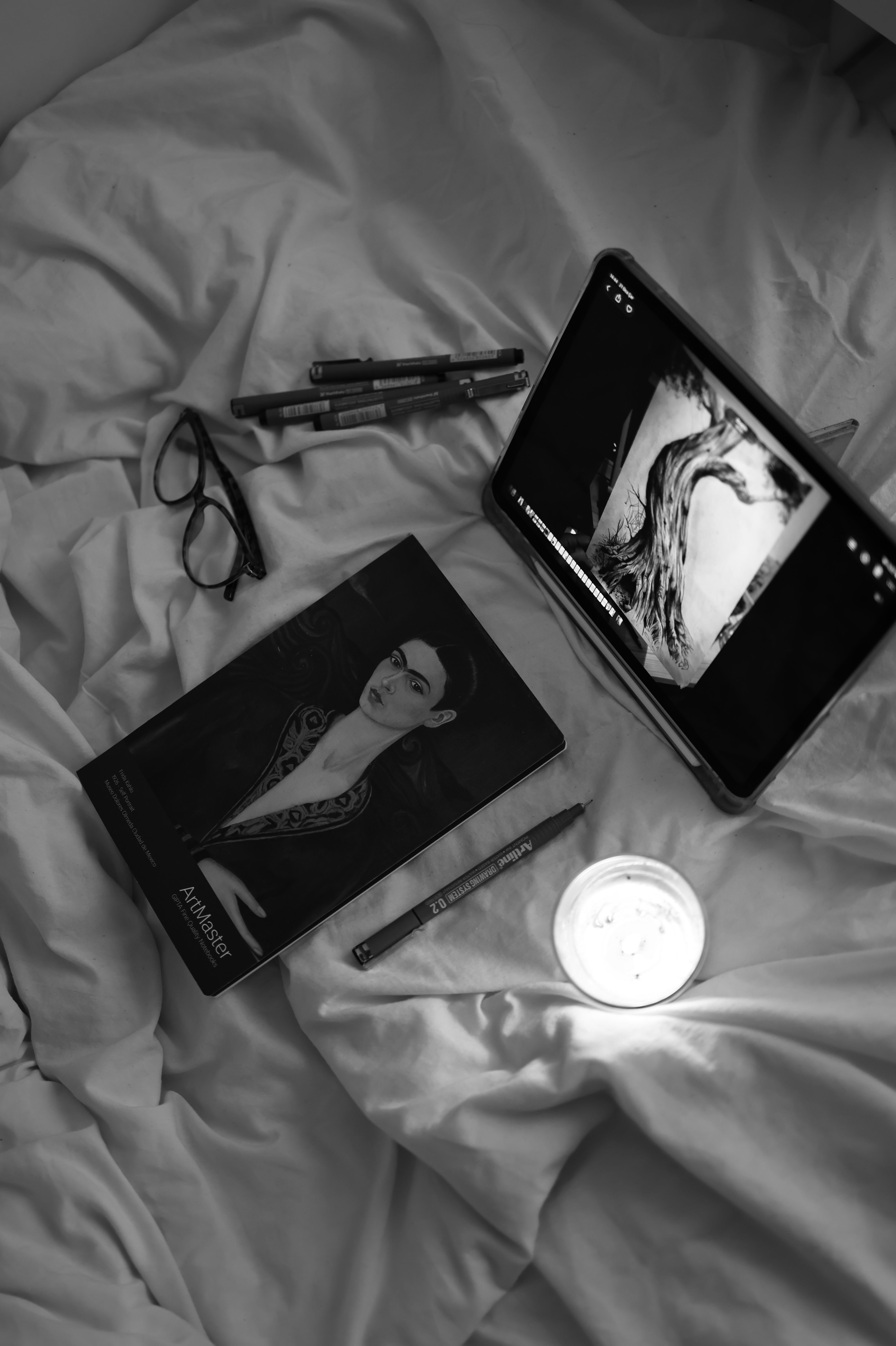 Black and white photo of a tablet, art book, pens, and candle on a bedspread.