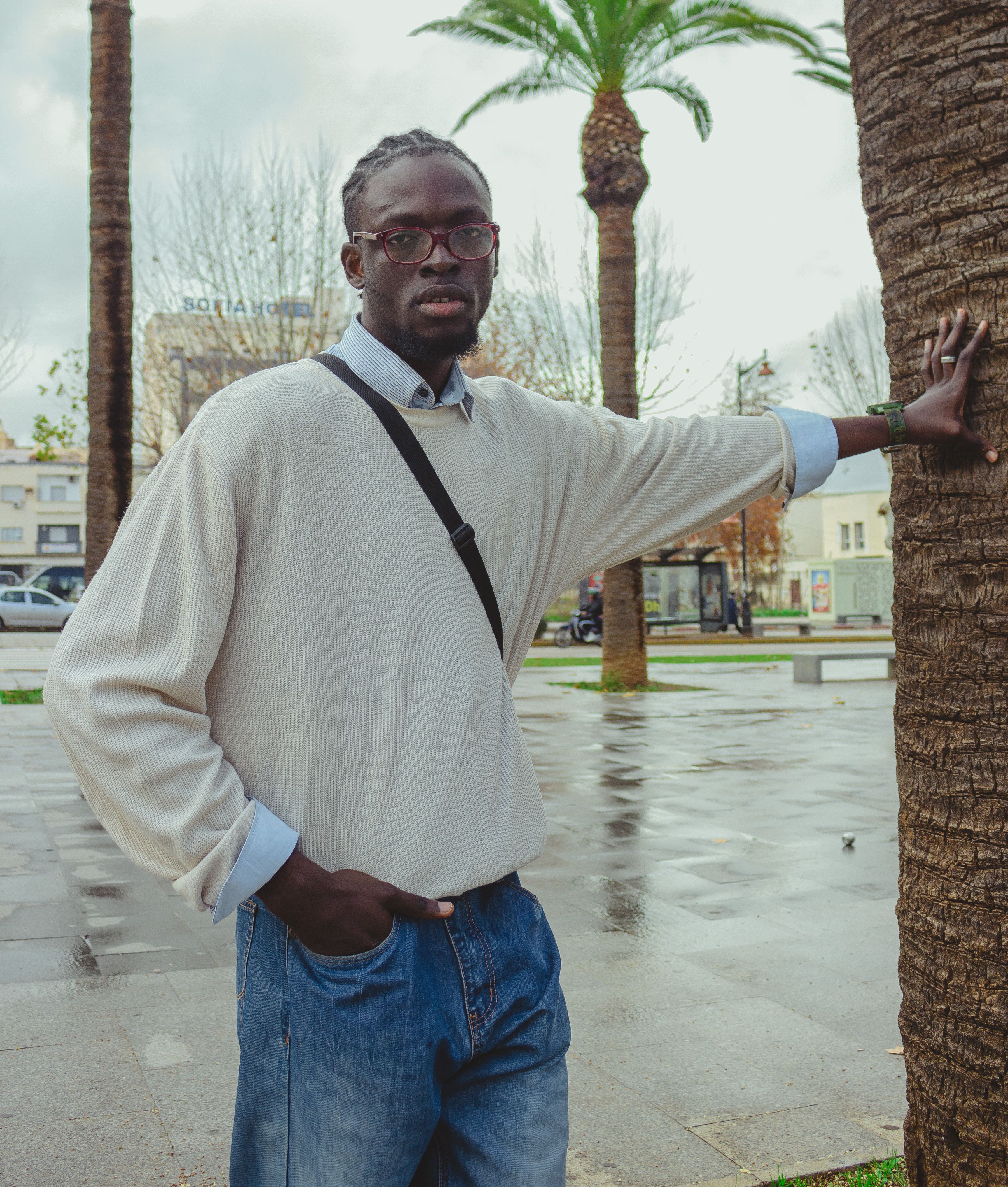 Fashionable adult male wearing glasses and casual attire, standing beside a palm tree in an urban park setting.