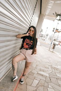 Young woman in stylish outfit poses on urban street with tattoo and sunglasses.