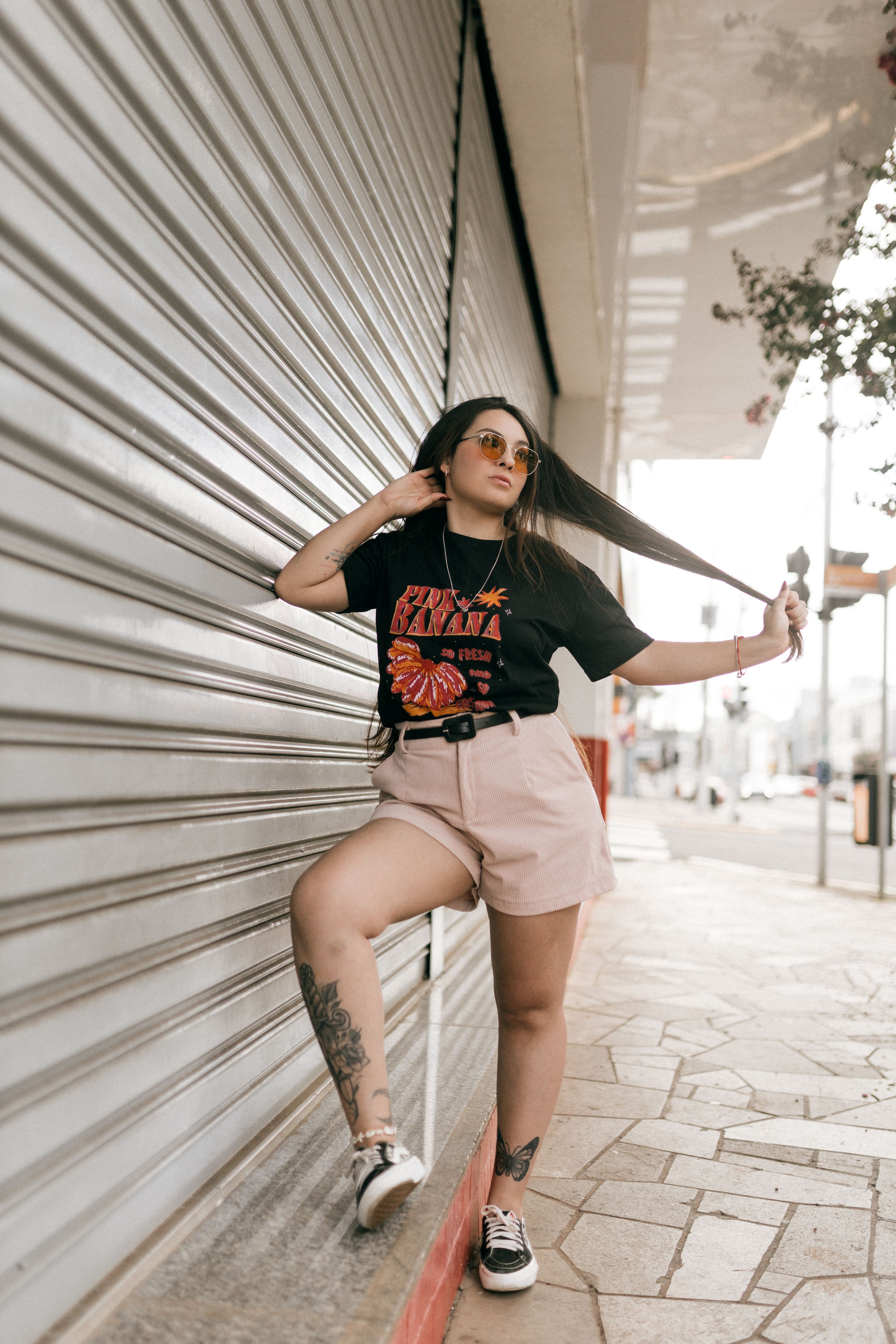 Mulher estilosa em traje urbano posando ao lado de uma porta de garagem na cidade, usando óculos de sol e shorts.
