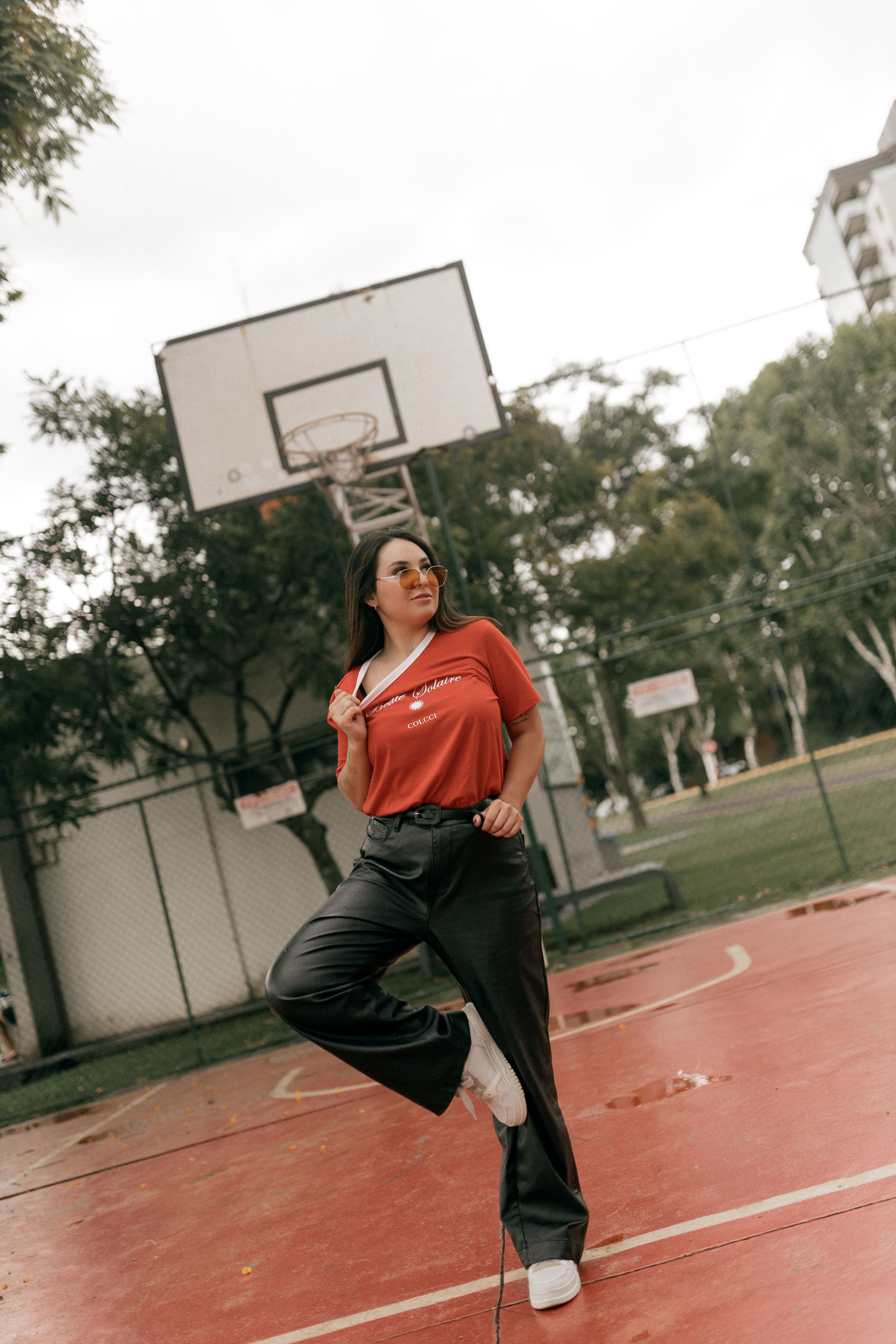 Confident woman in red shirt poses on an outdoor basketball court, showcasing urban style.