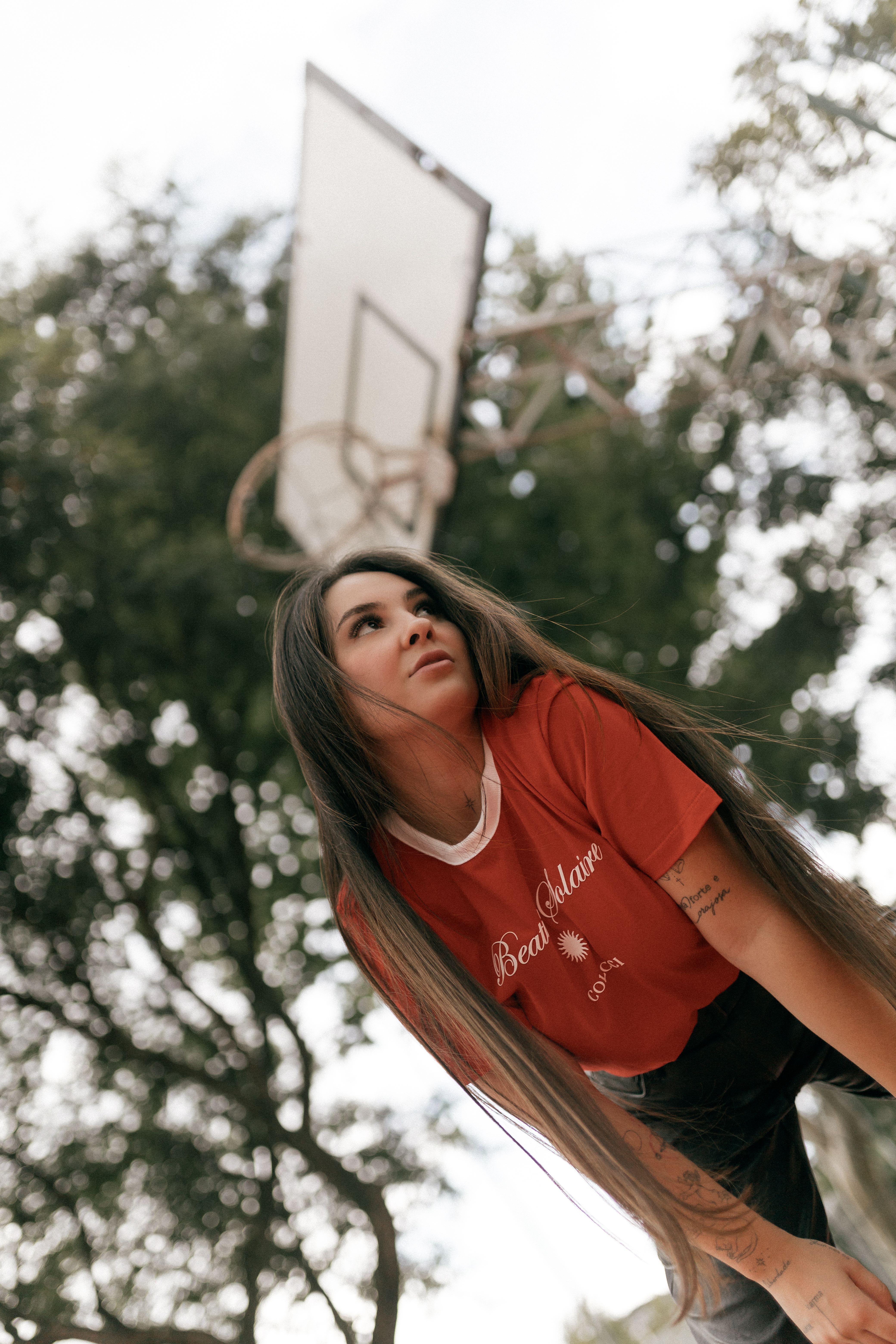 Woman in red shirt posed under basketball hoop outdoors with trees.