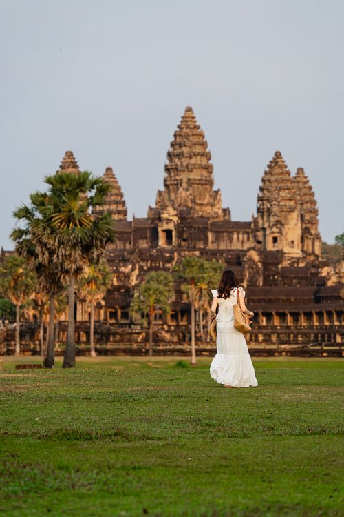 Free A woman in a white dress walks towards Angkor Wat, Cambodia, amidst lush greenery during a serene evening. Stock Photo