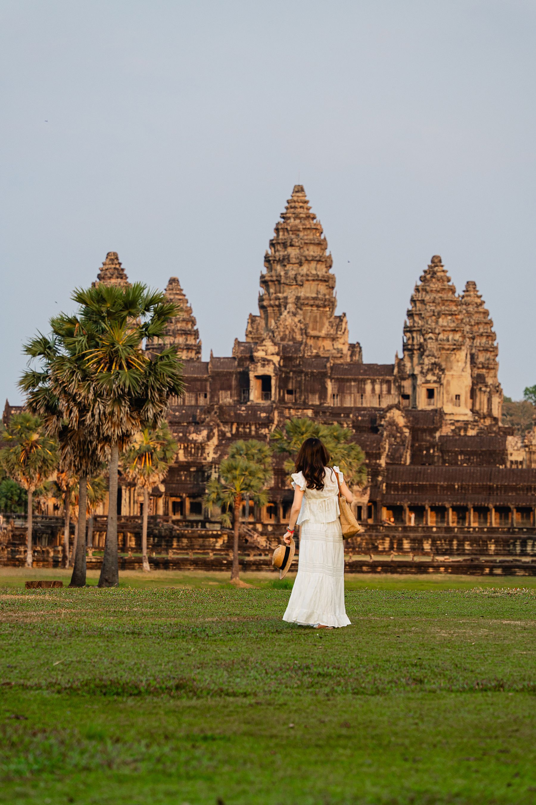 Traveler crossing green grounds toward the complete five-tower profile of Angkor Wat