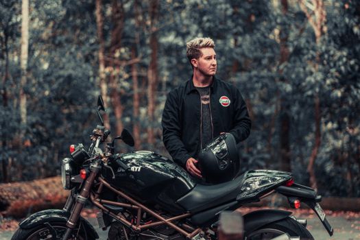 A man standing with a Ducati motorcycle in a forest setting, exuding a retro aesthetic.