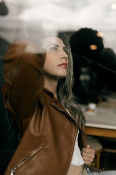 Young woman in a brown leather jacket seen through glass indoors with soft lighting.