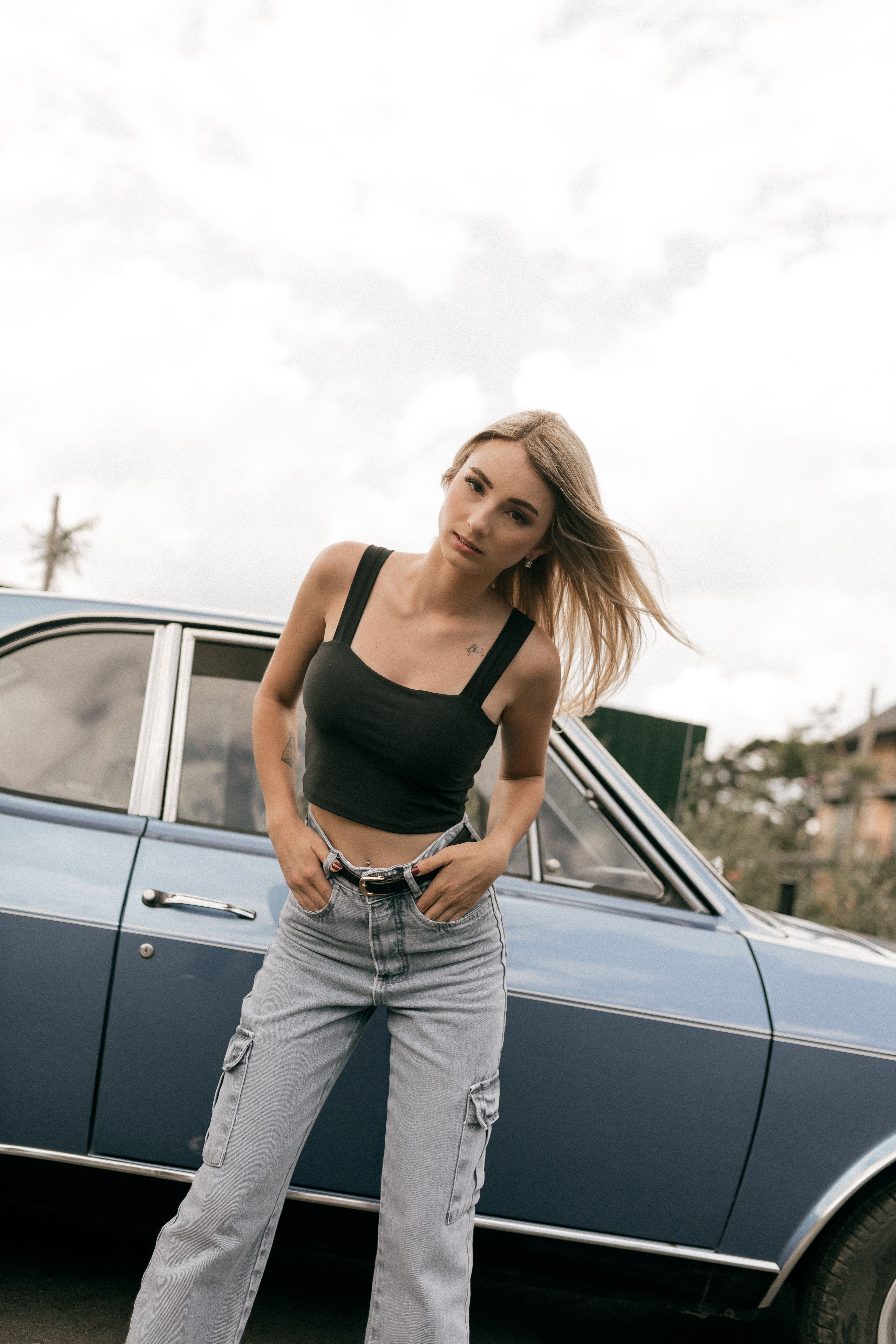 Trendy woman in casual outfit standing confidently by a vintage blue car on a cloudy day.