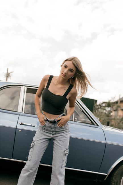 Trendy woman in casual outfit standing confidently by a vintage blue car on a cloudy day.