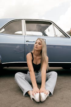 Stylish young woman sits casually in front of a vintage blue car on a sunny day.