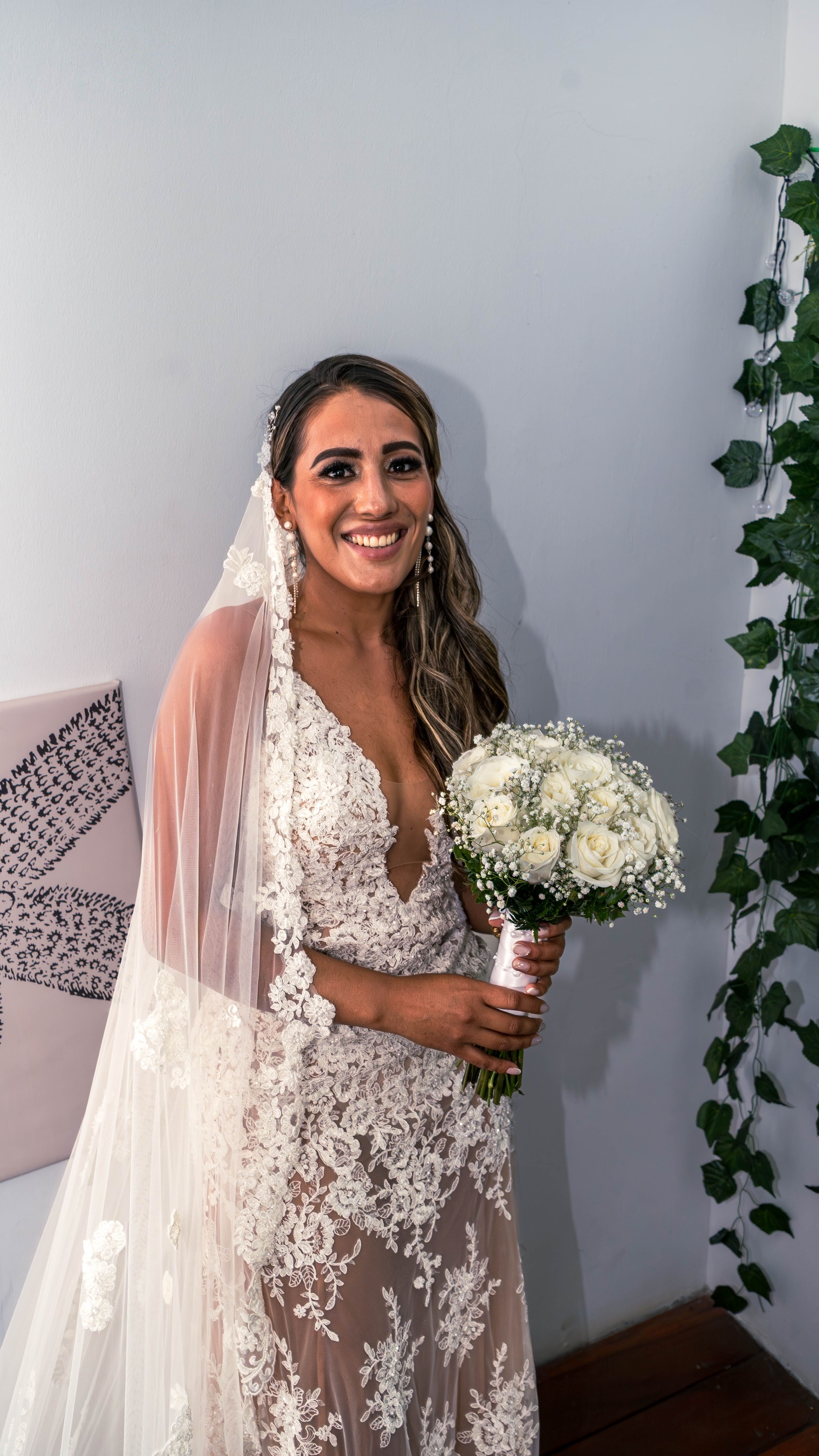 Beautiful bride in lace gown with a bouquet, smiling indoors against a decorated wall.