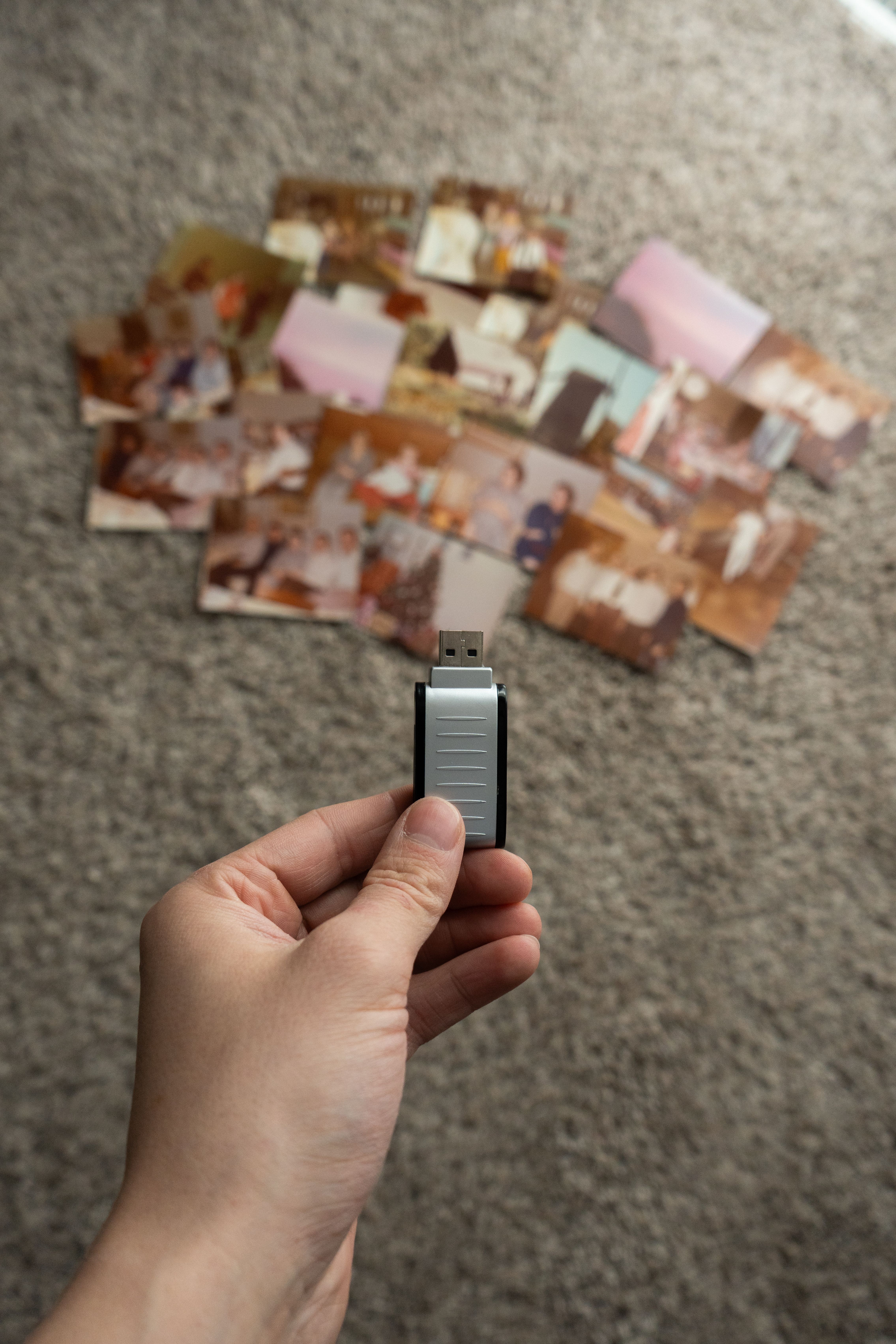 Hand holding USB drive with vintage photos scattered on carpet, symbolizing digital archiving.