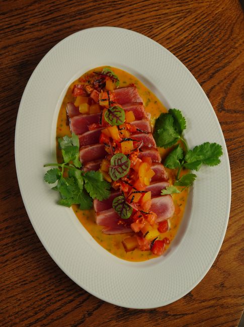 Sliced seared tuna tataki fanned on a plate, showing a thin gray-brown seared rim around a raw red center, garnished with scallions and sesame seeds
