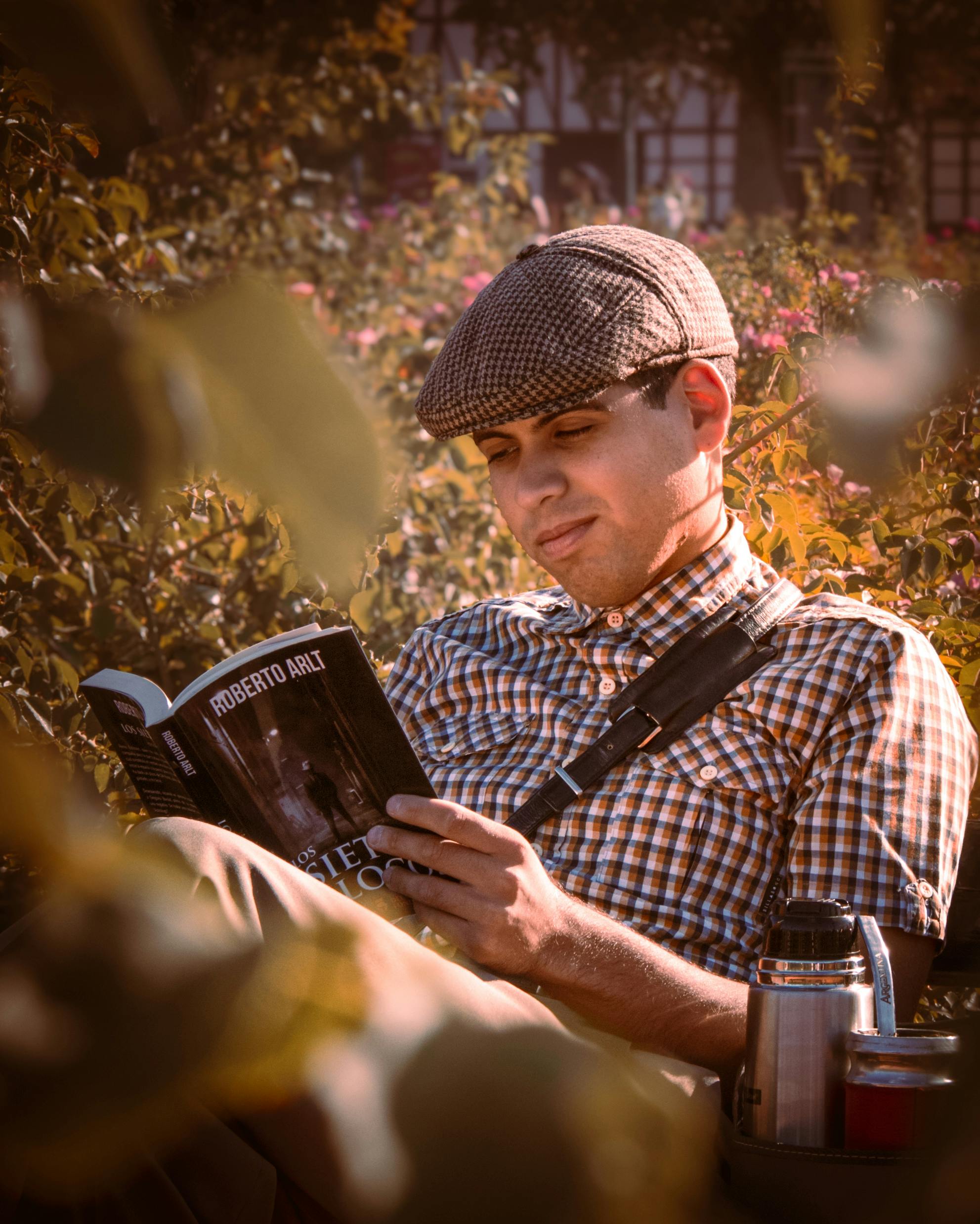A man in checkered shirt reading a book in a garden setting with autumn vibes.