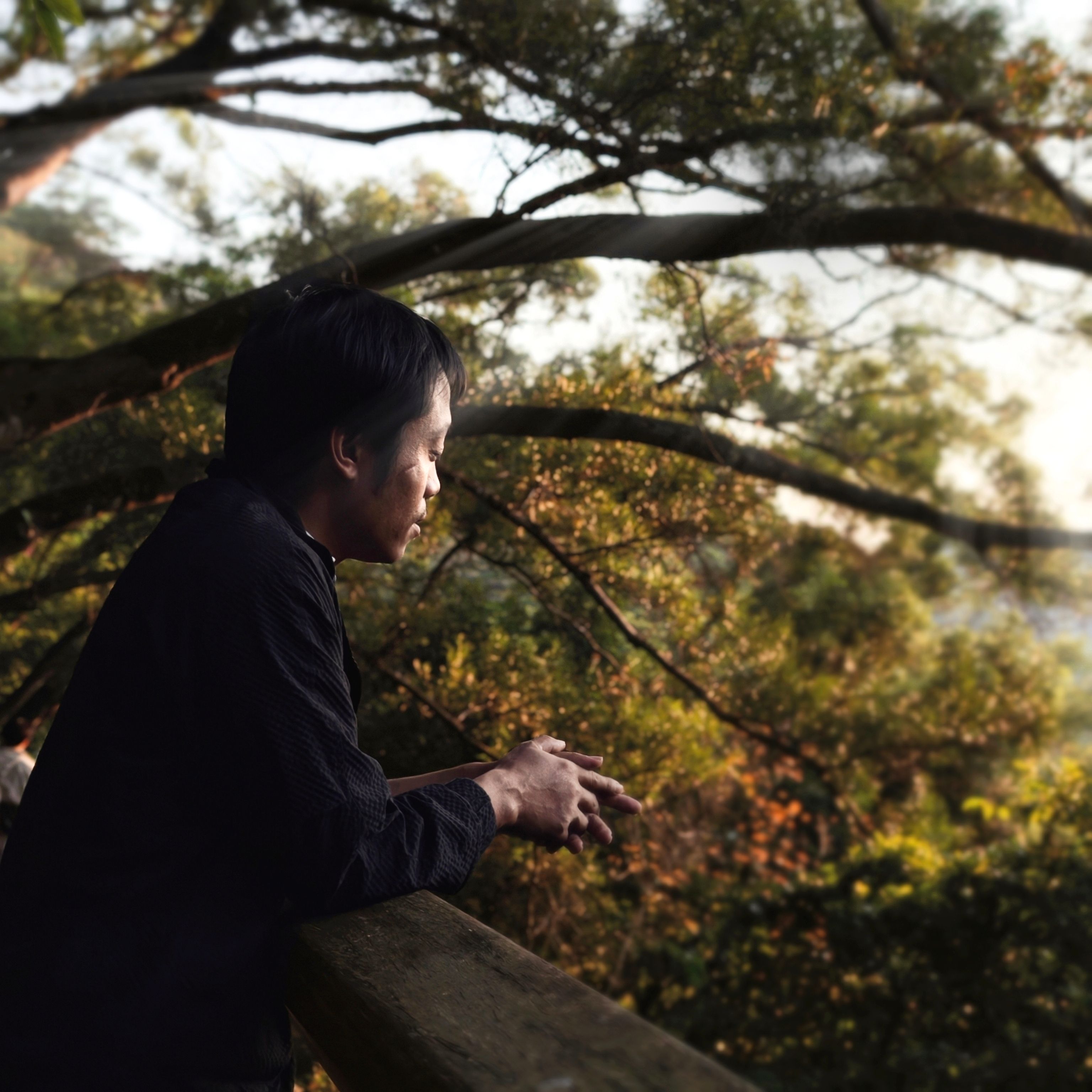 A man leans on a wooden railing, lost in thought, surrounded by lush greenery during sunset.