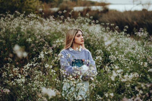 Young woman stands thoughtfully among wildflowers, holding a book in serene outdoor setting.