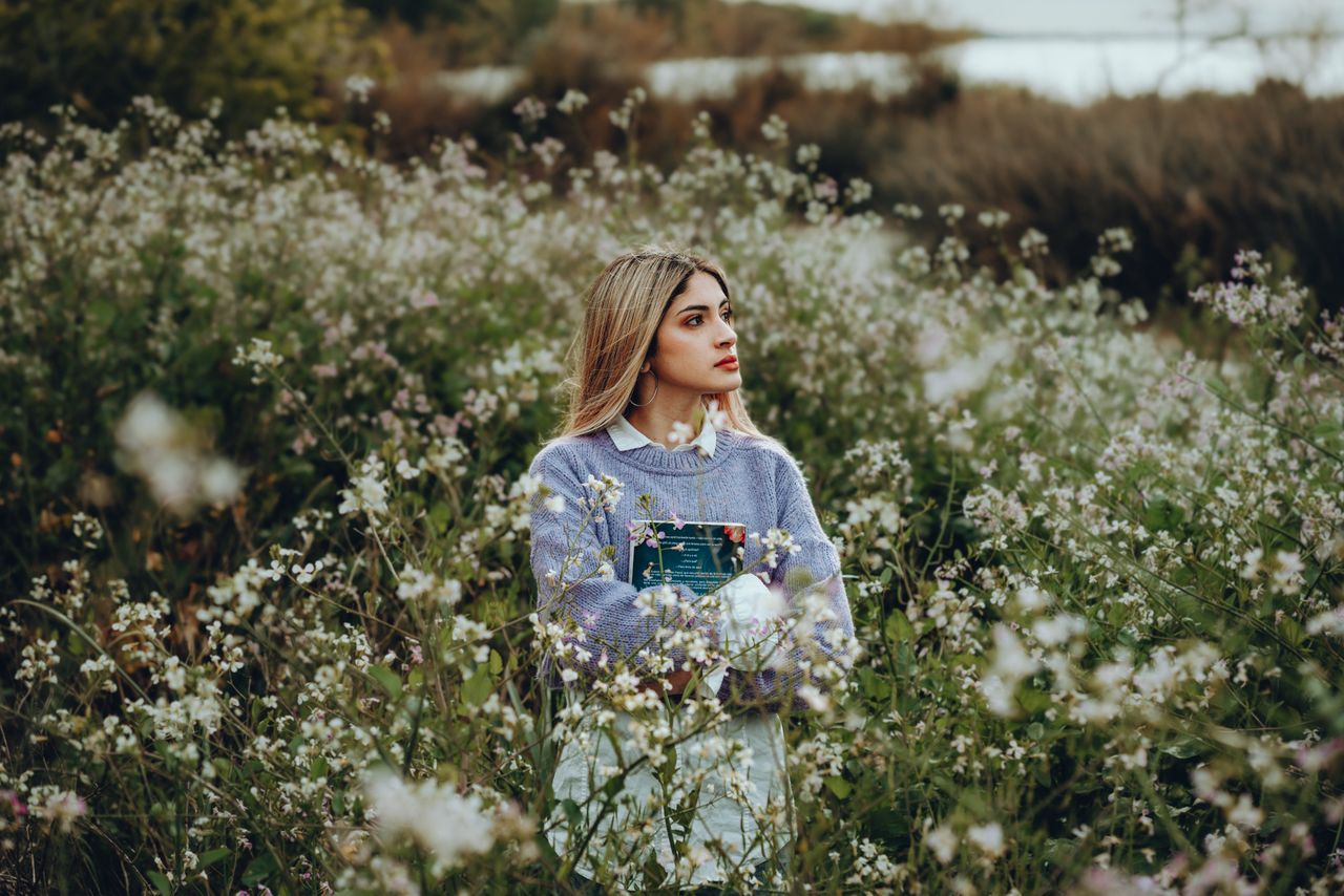 young woman stands thoughtfully among wildflowers holding a book in se