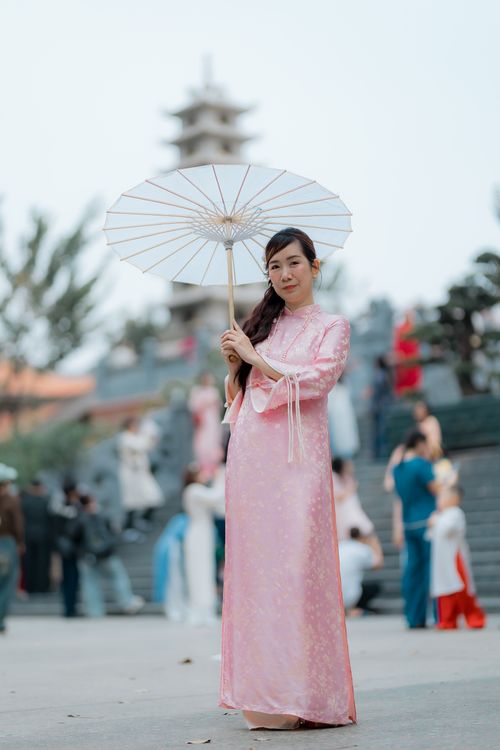 Free A woman in a pink traditional dress holding an umbrella, standing outdoors with a pagoda in the background. Stock Photo