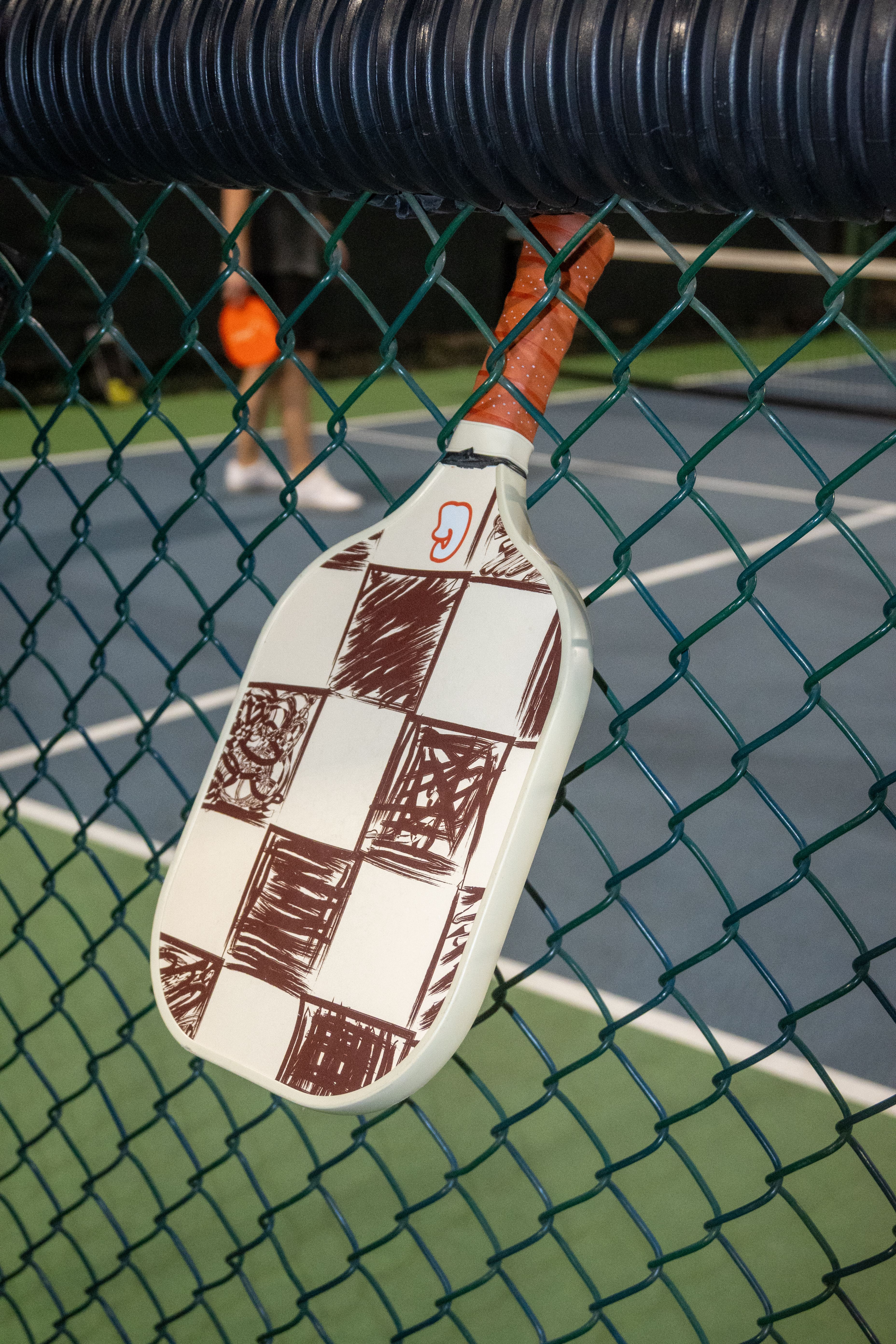 Close-up of a pickleball paddle with a checkered design hanging on a court fence at night.