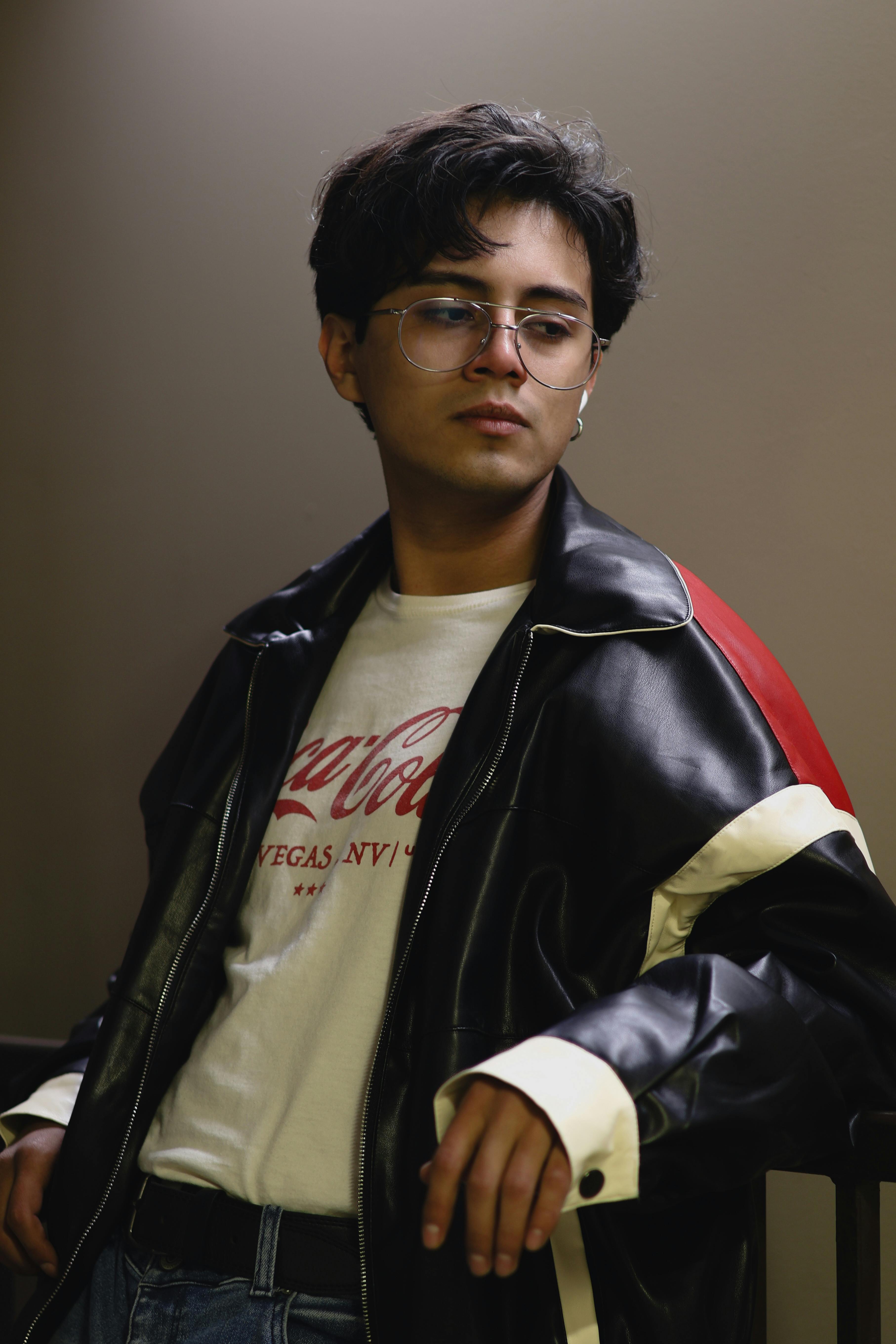 Fashionable teenager with glasses wearing a leather jacket, posing indoors with a moody expression.