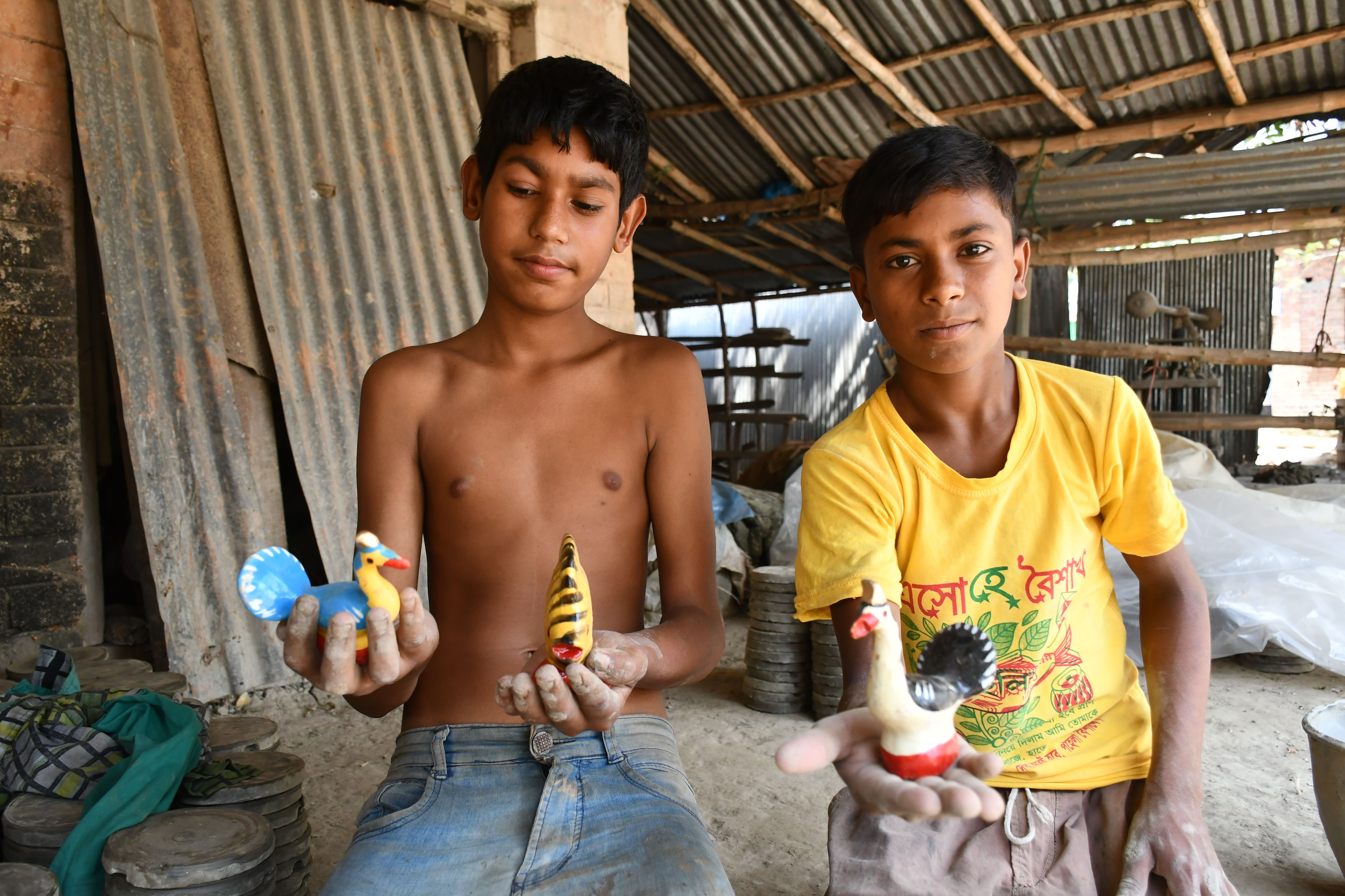 Two boys showcasing colorful handmade clay toys in a rustic workshop in Rangpur, Bangladesh.